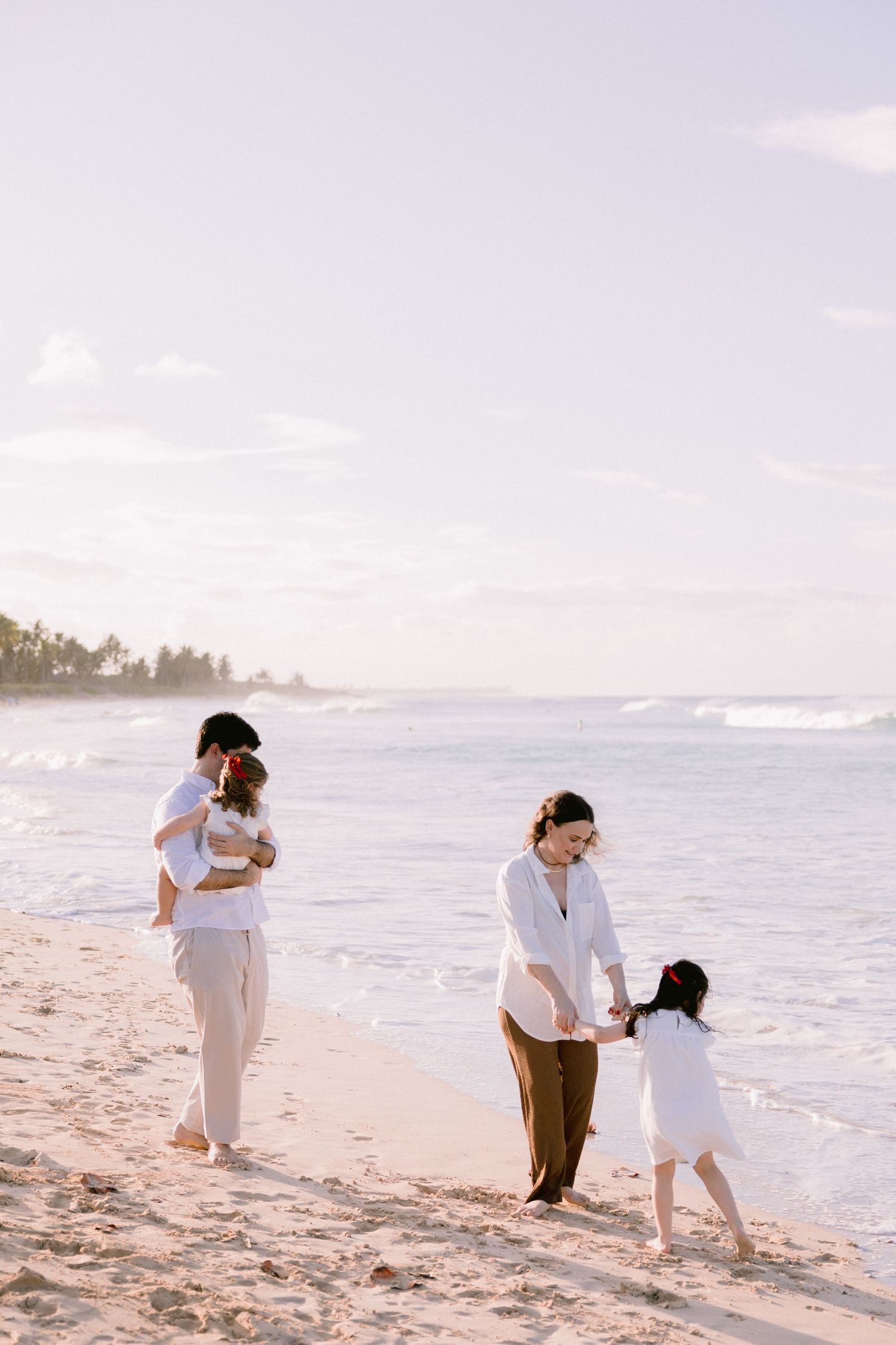 Family in white outfits enjoying a sunny beach walk, parents playing with young children on sandy shore near ocean waves.