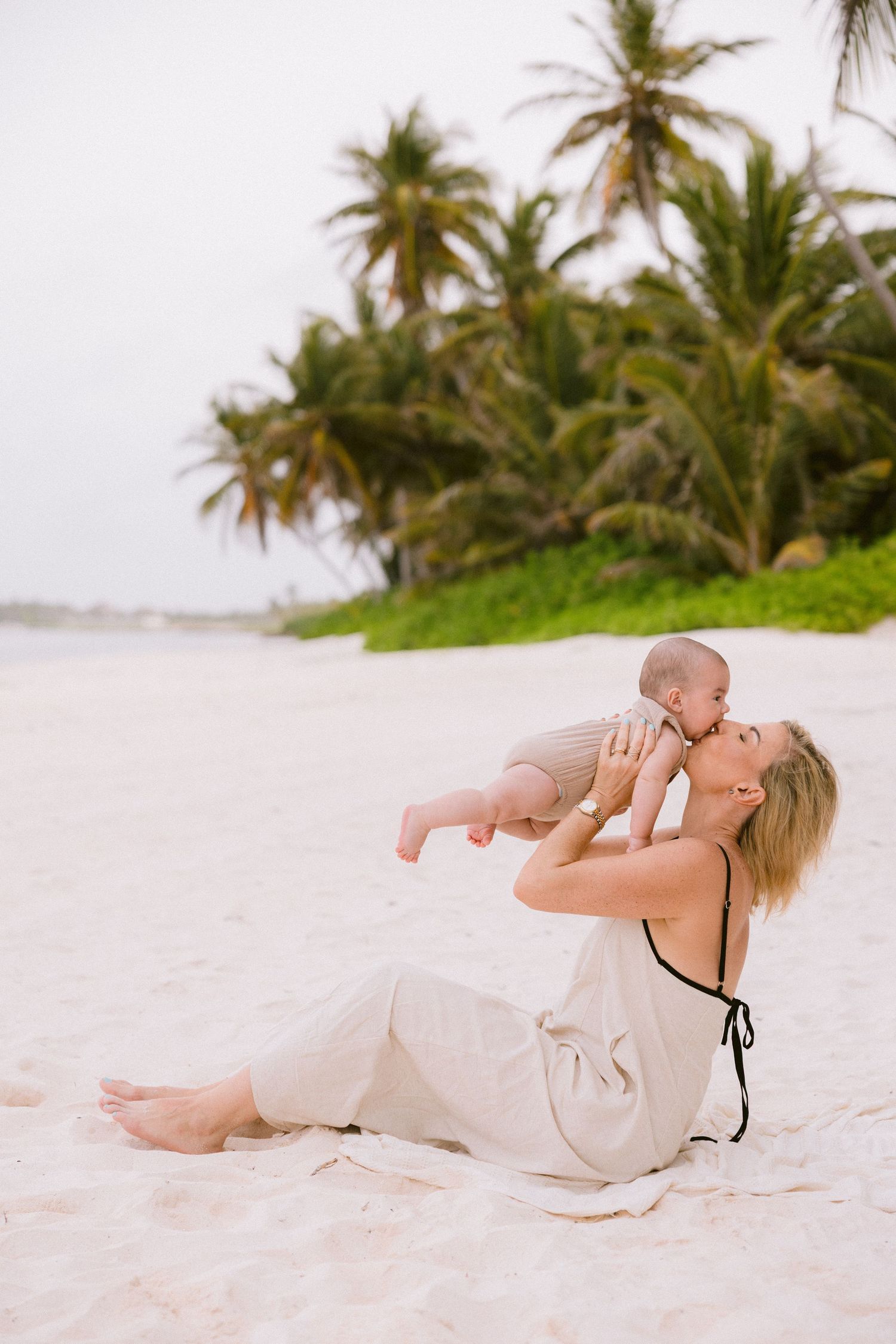 Mother and baby playing together on sandy beach with palm trees in background.