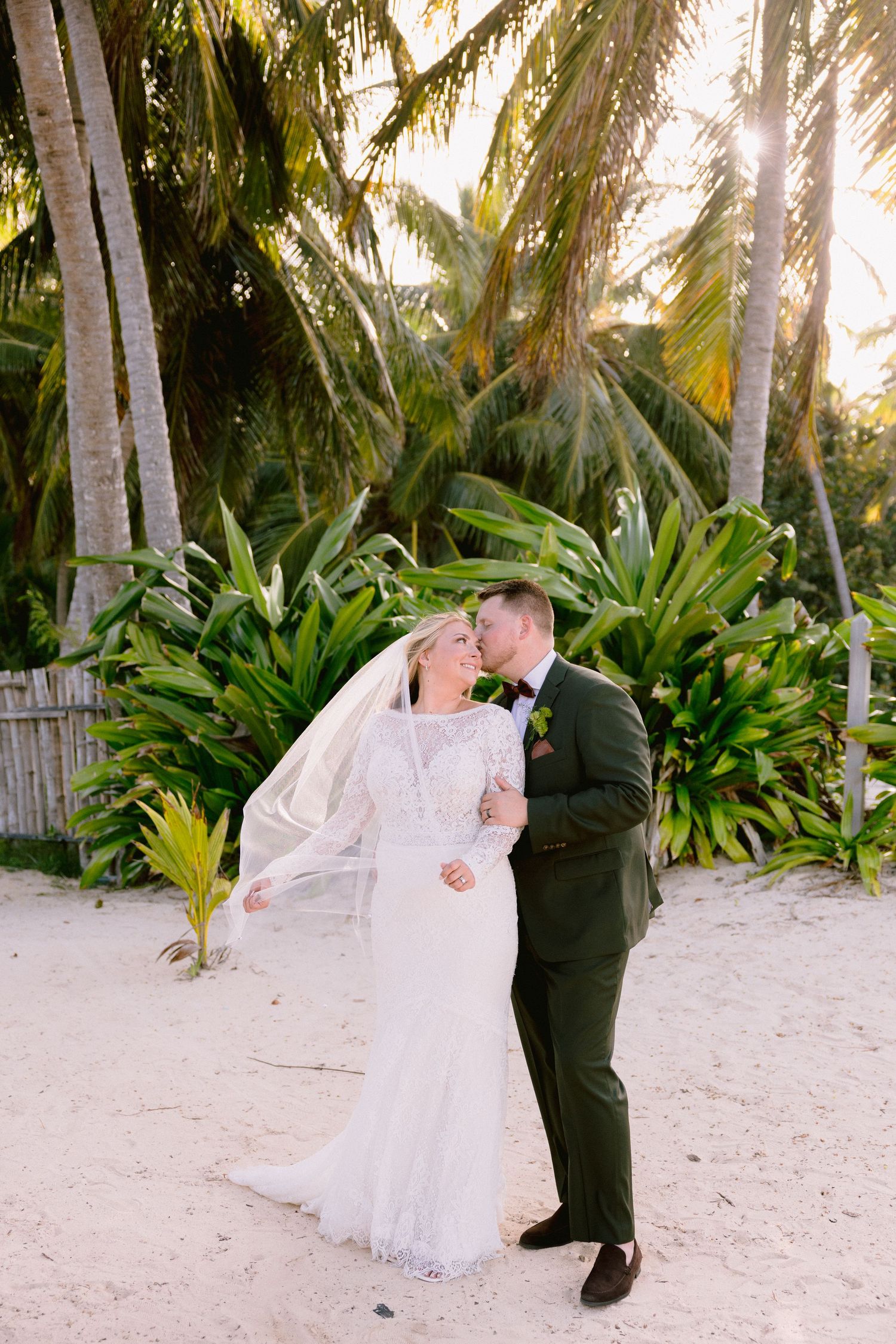 Bride and groom kissing on sandy beach surrounded by tropical palm trees and lush greenery in golden sunset light.
