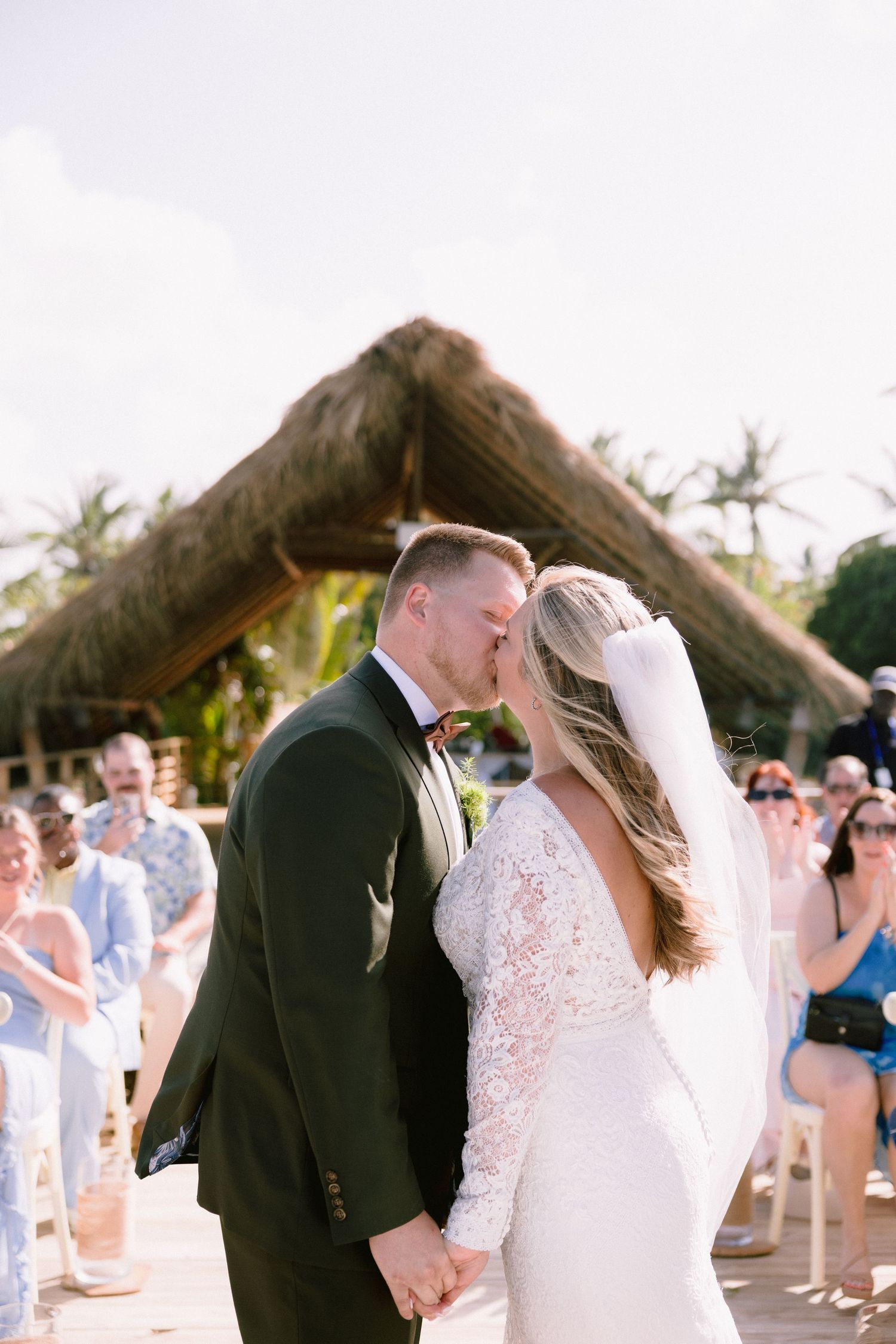 Bride and groom share first kiss at outdoor tropical wedding ceremony with guests applauding in background.