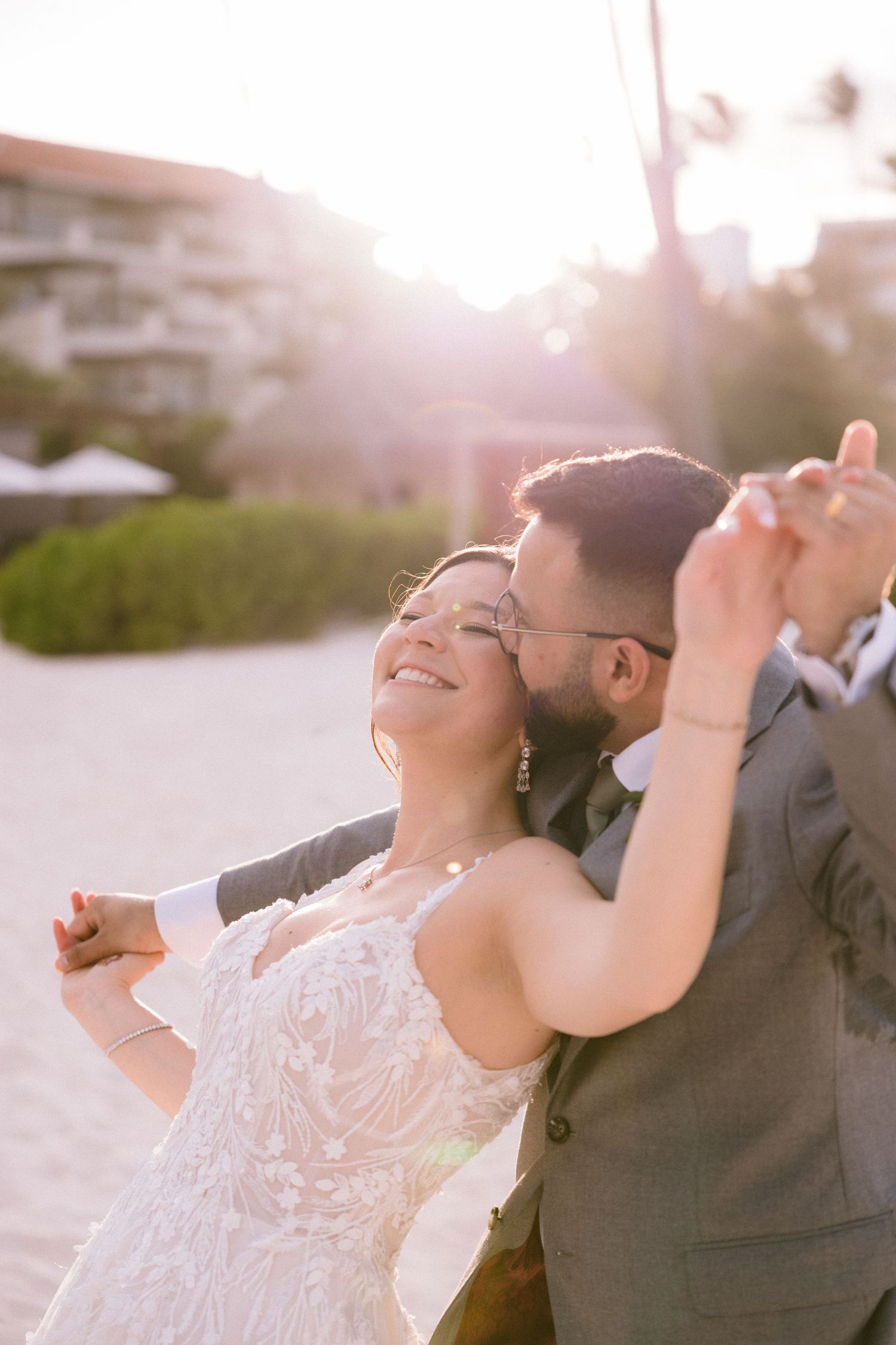 A couple in wedding attire dance joyfully on a sunlit beach during golden hour.