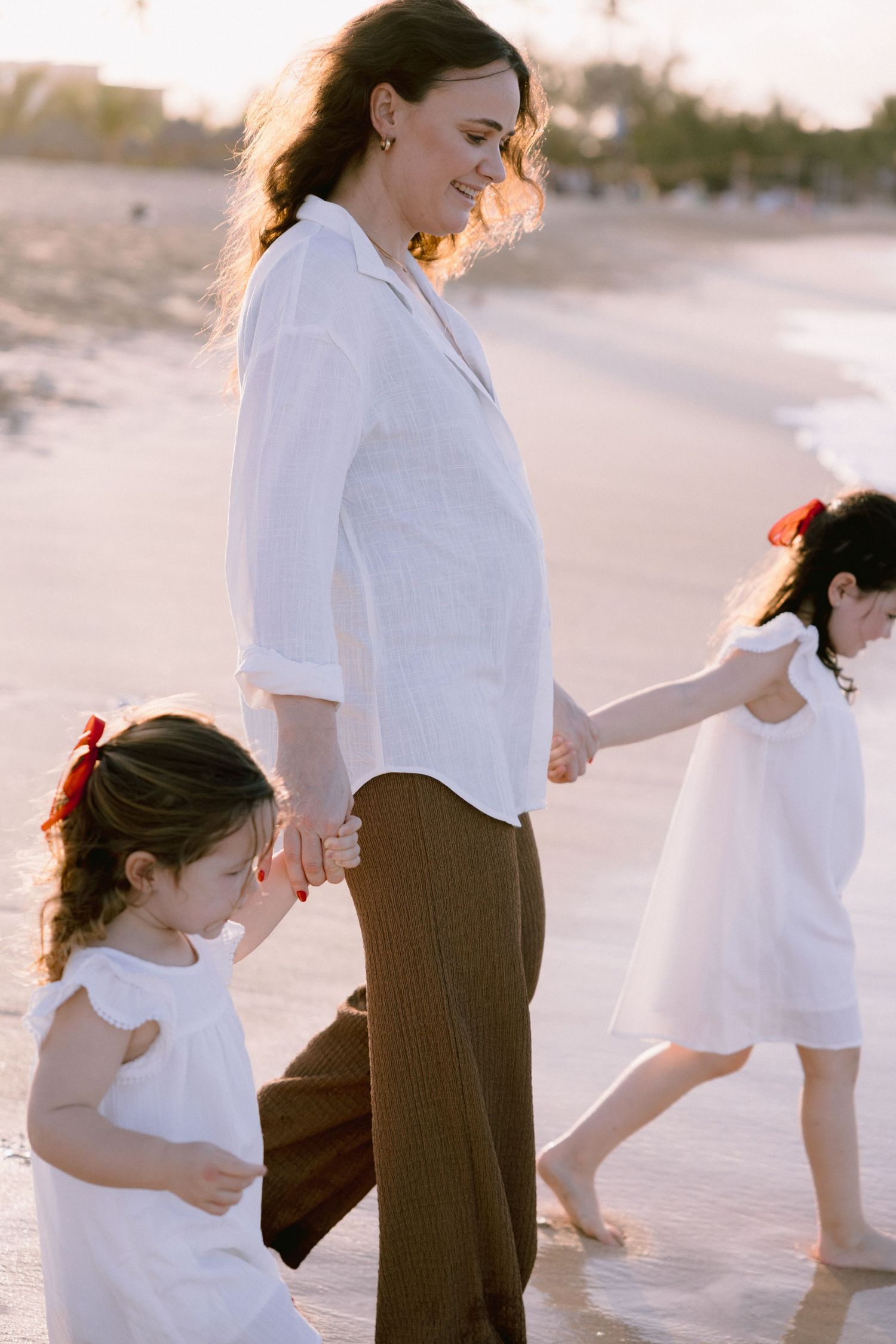A family walks together on a sunny beach at sunset, wearing white and brown casual clothing.