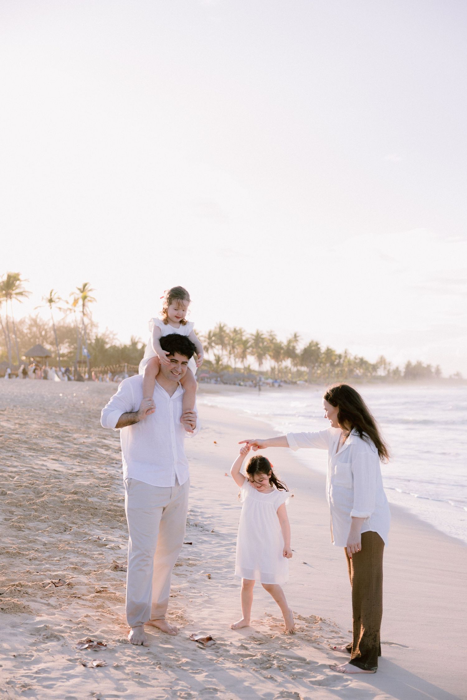 Family in white outfits walking together on a sandy beach at sunset with palm trees in the background.