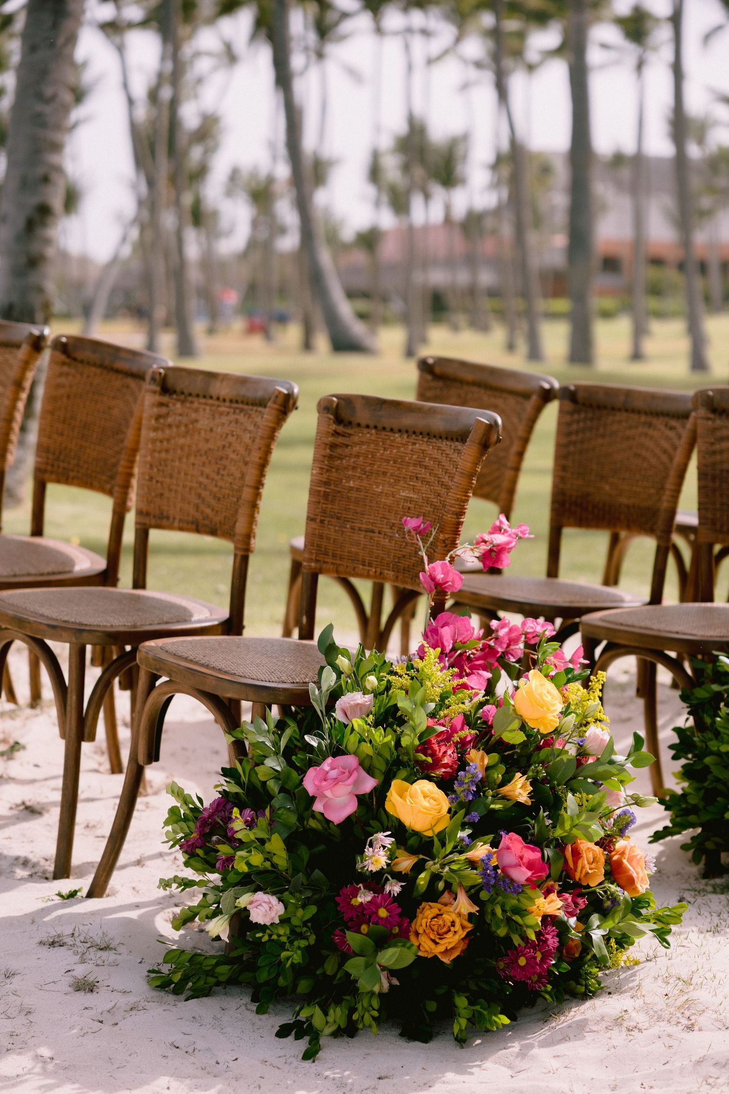Brown rattan chairs arranged for an outdoor ceremony with vibrant pink and yellow floral arrangements lining the aisle.