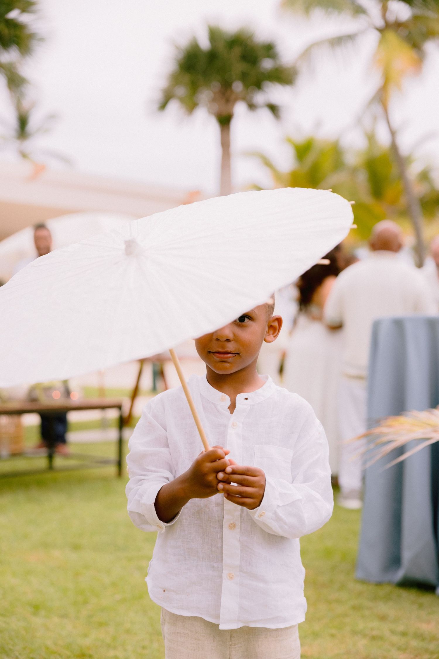 Young boy in a white linen shirt holds a white paper parasol at an outdoor tropical wedding with palm trees and guests in the background.