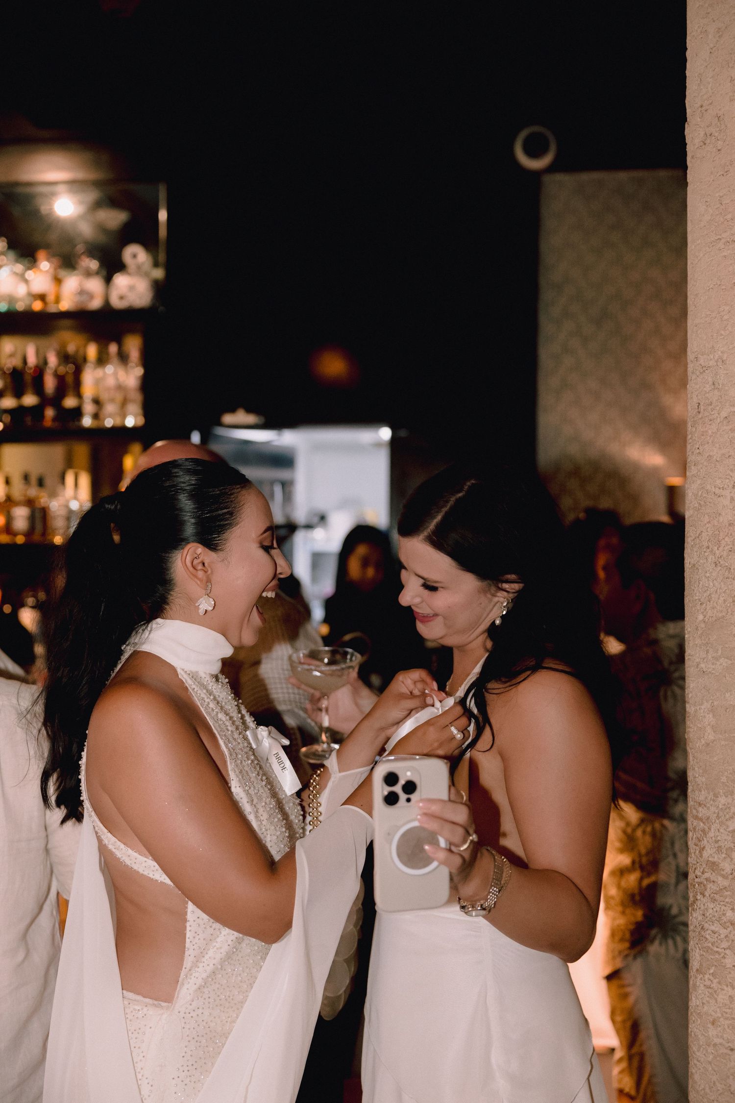 Two women in white dresses laugh together at a dimly lit bar, one holding a coupe glass and the other a smartphone.