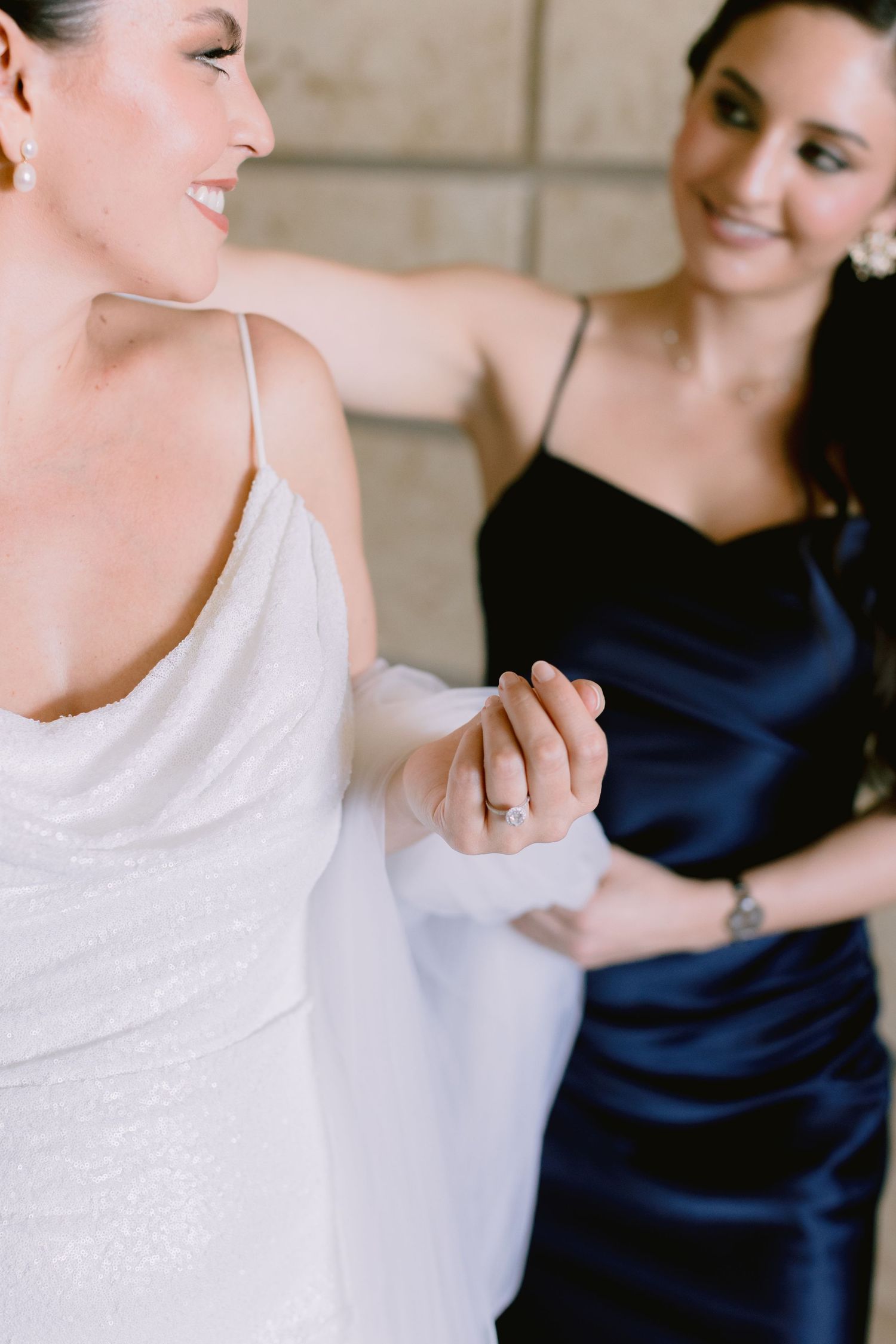 Bride in white gown holds hands with bridesmaid in navy satin dress during wedding preparation.