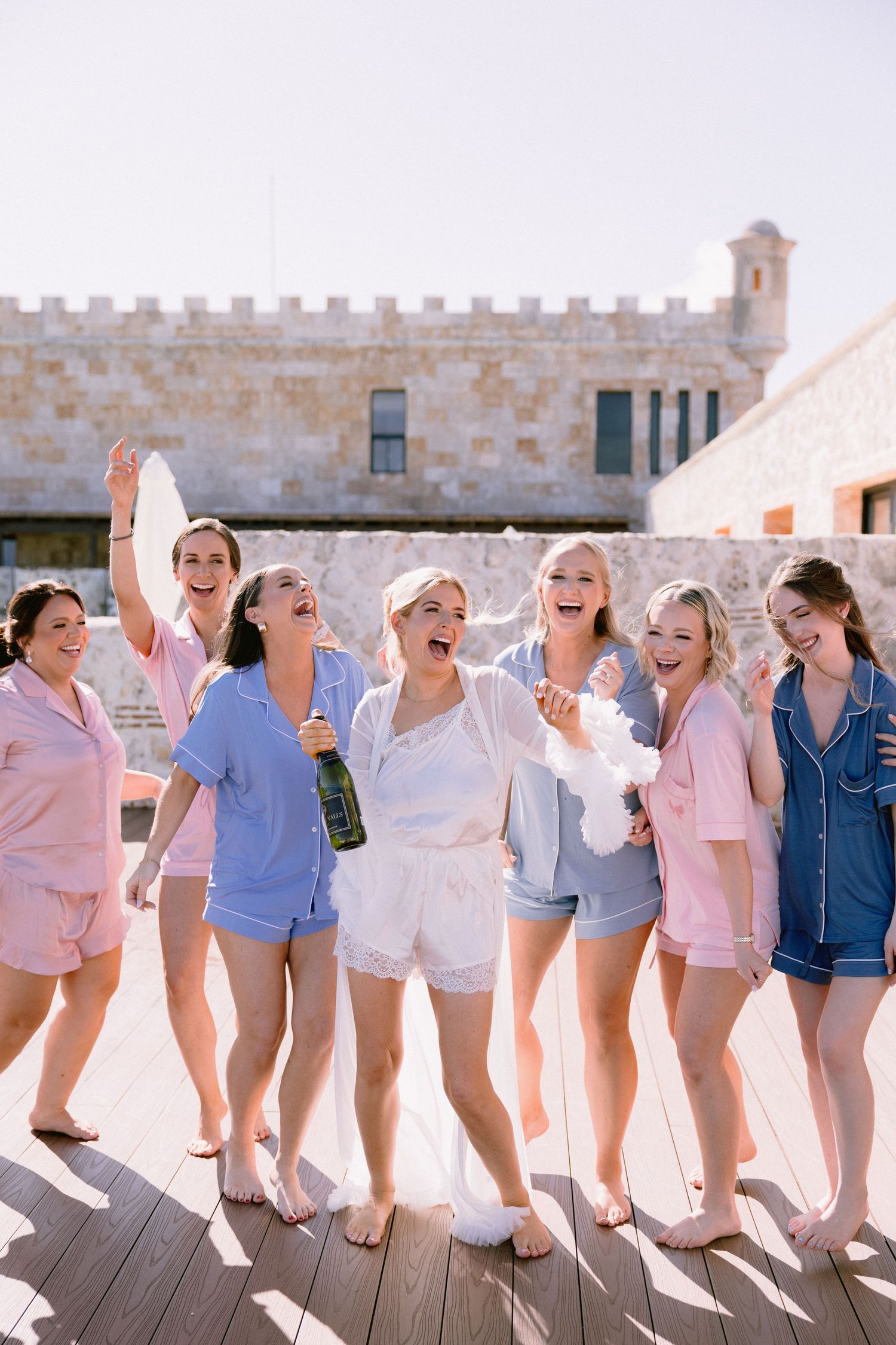 Bridesmaids in colorful pajama sets celebrate with champagne outdoors near a stone building on a sunny day.