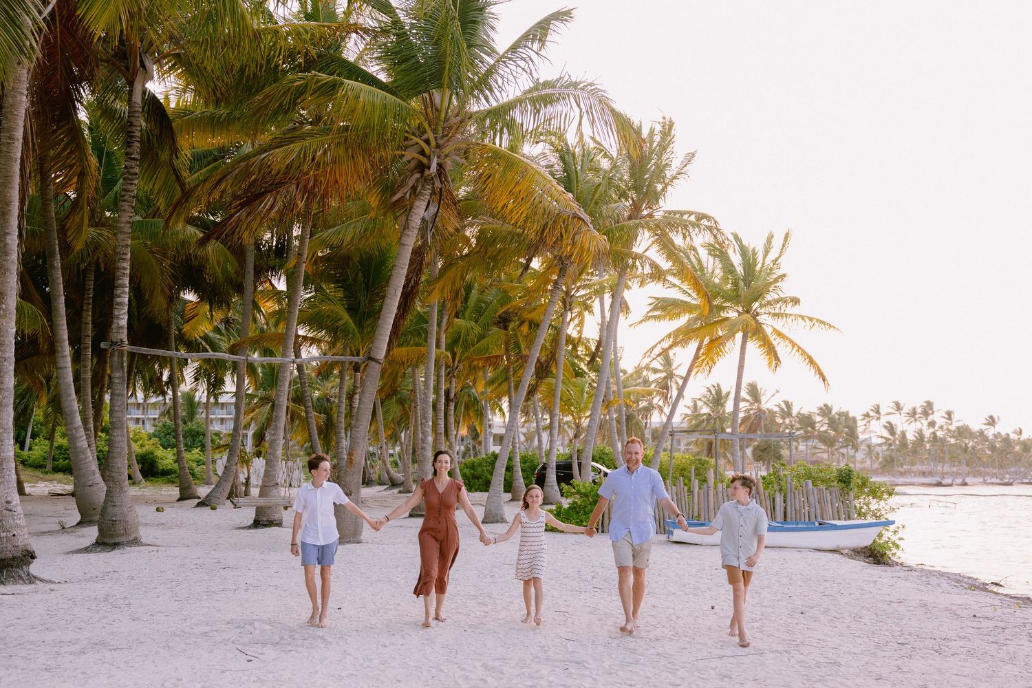 Family of four walking on a tropical beach with tall palm trees and white sand at sunset.