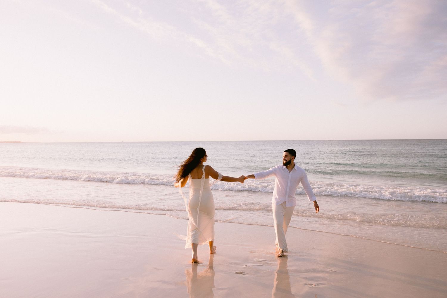 Couple in white outfits holding hands walking on a sandy beach at sunset with gentle ocean waves.