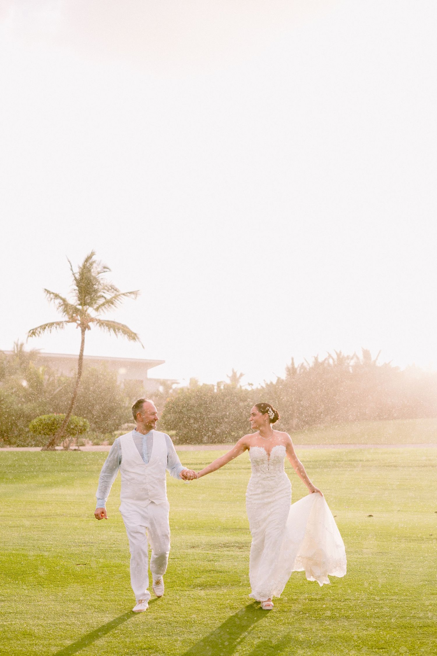 Bride and groom walk hand-in-hand on lush green lawn at sunset, palm trees and warm golden light in background.