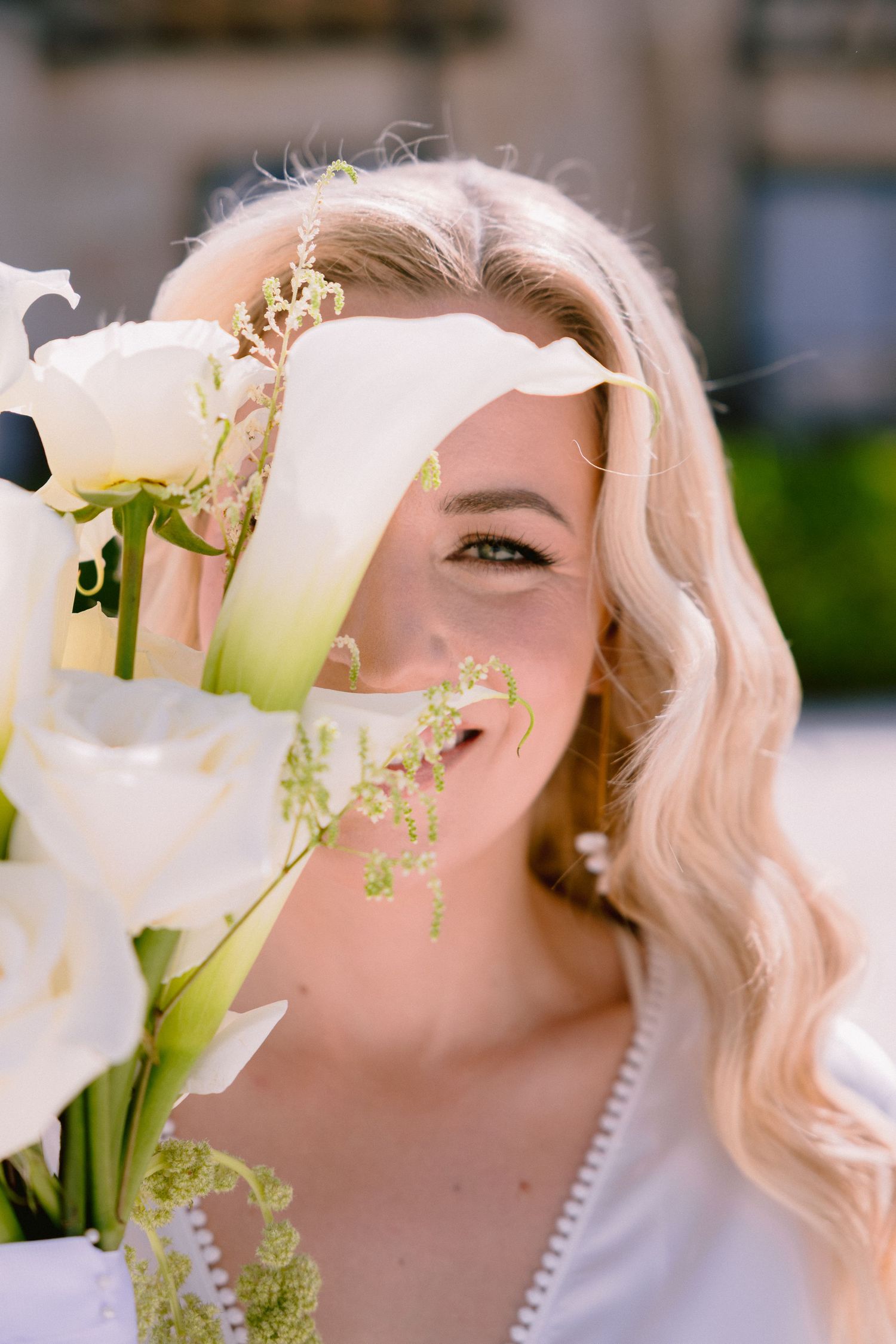 Smiling blonde bride holding white calla lily bouquet covering her face, outdoor wedding portrait.