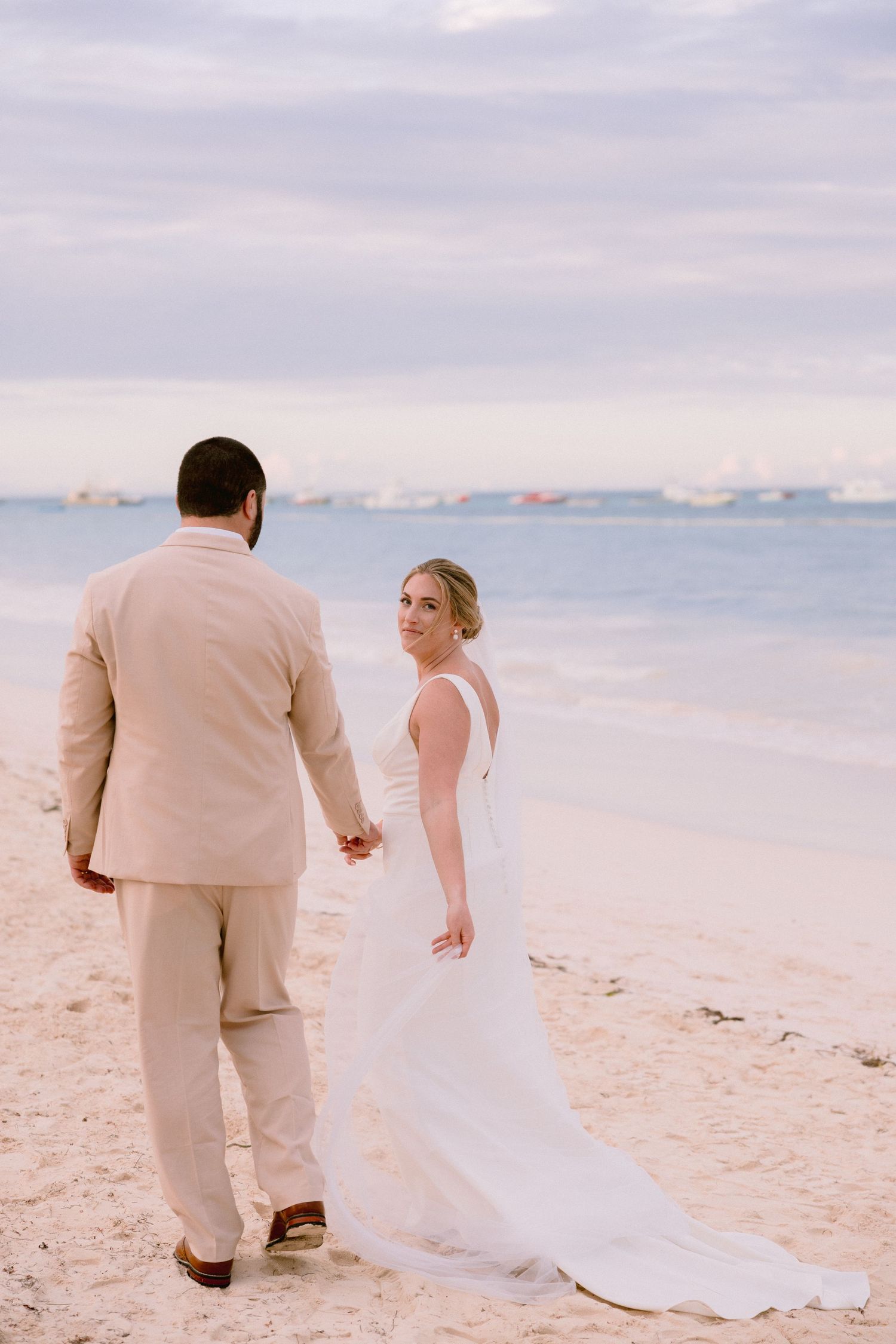 Bride in white gown and groom in tan suit hold hands on a sandy beach with calm ocean waters in the background.