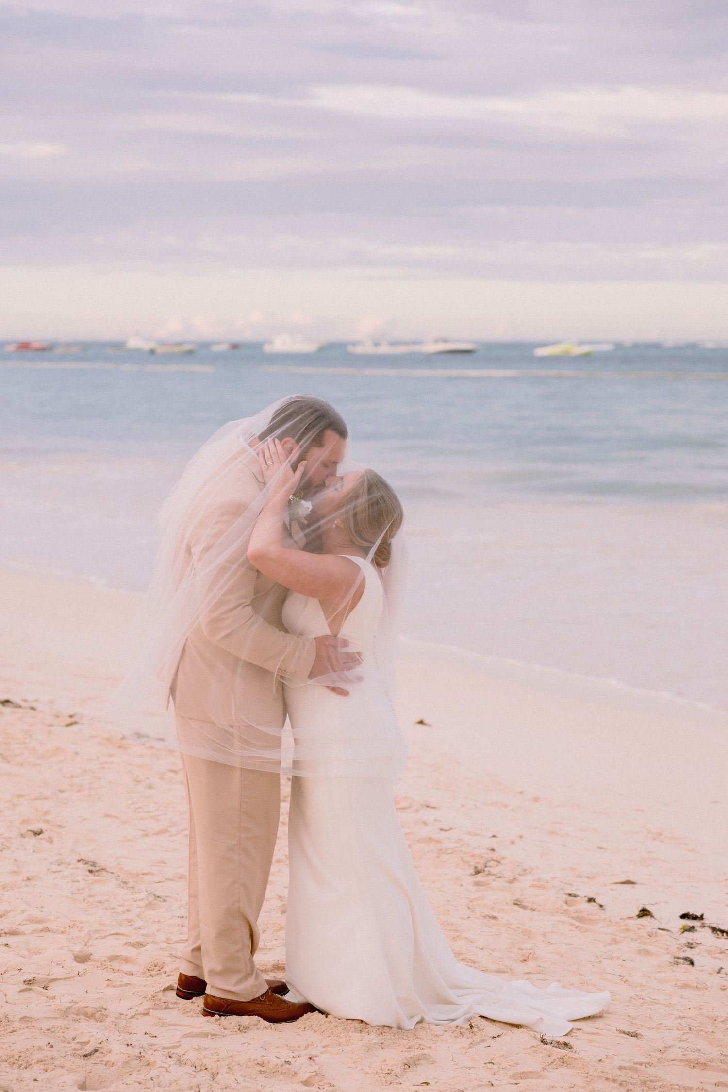 Bride and groom kissing under a veil on a sandy beach with calm turquoise ocean waters in the background.