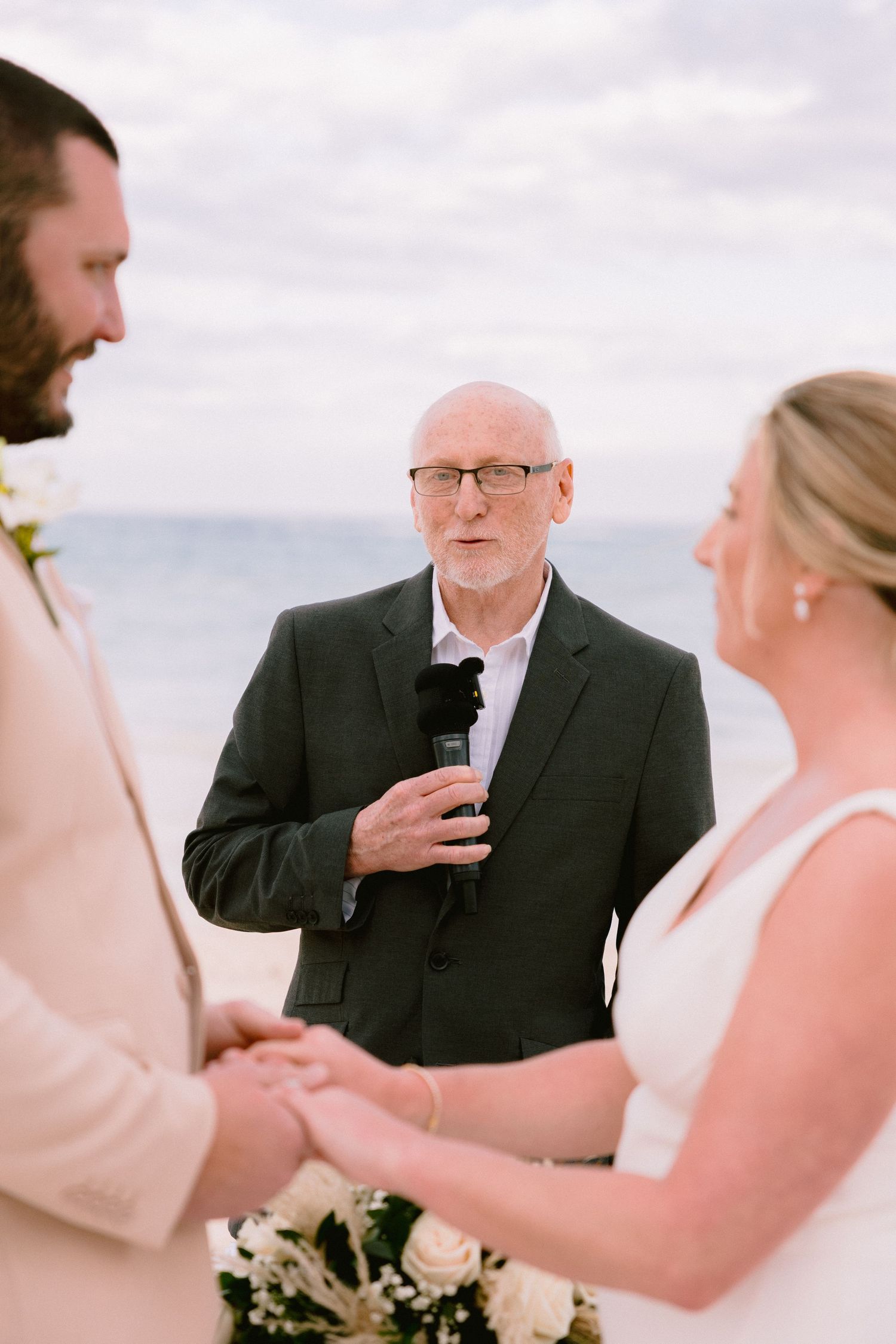 Officiant speaks at beach wedding ceremony as couple holds hands with ocean backdrop.