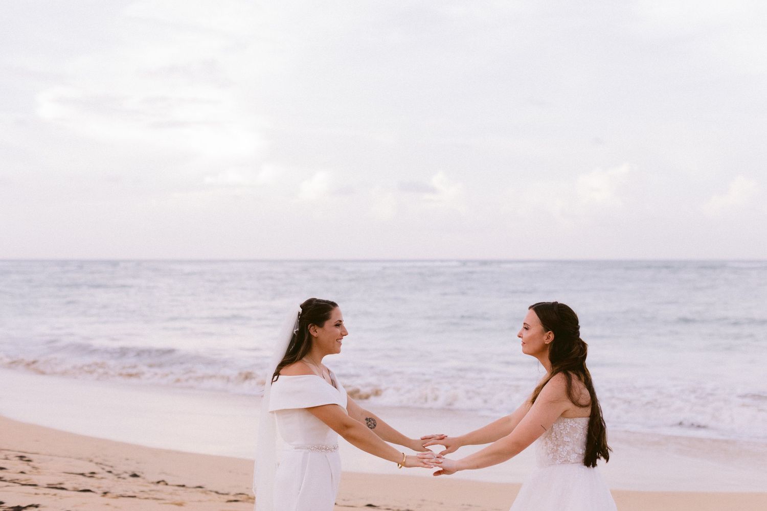 Two brides in white dresses hold hands on a sandy beach with ocean waves in the background.