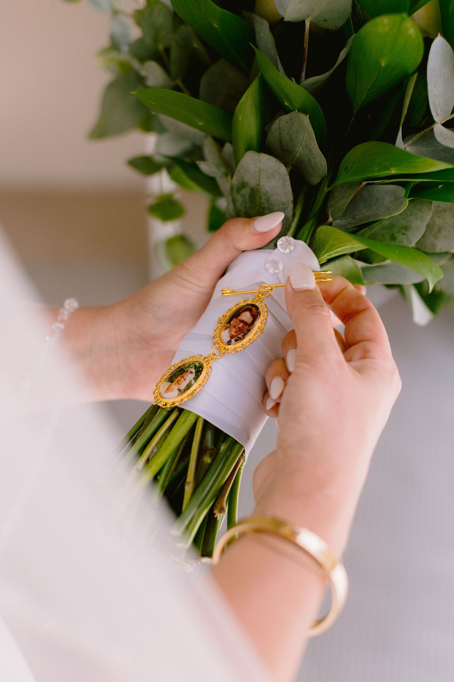 Bride holding wedding bouquet with gold locket photo charms tied with white ribbon, wearing gold bracelet.