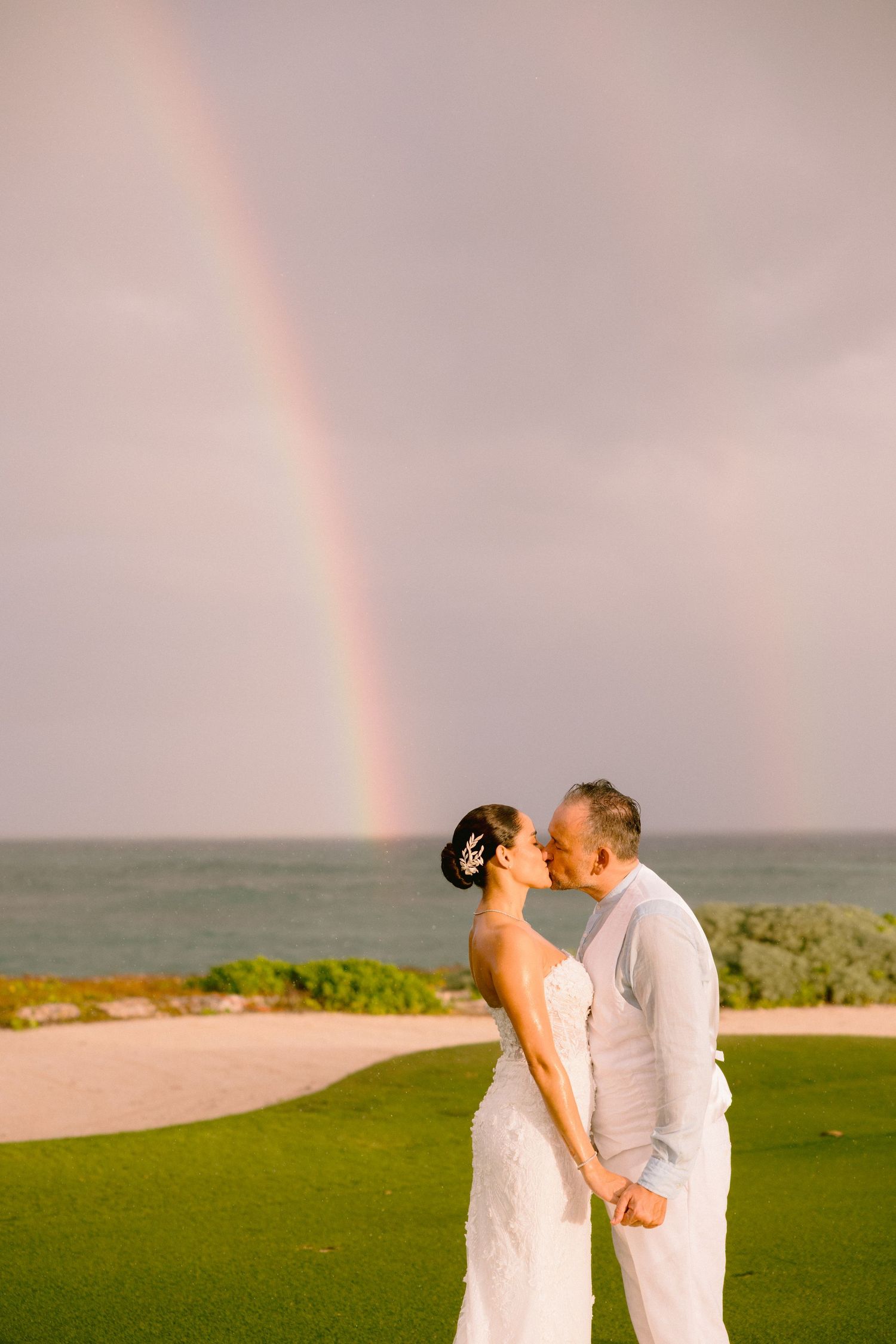 A couple shares a romantic kiss on a beach with a vibrant rainbow arching across the sky in the background.