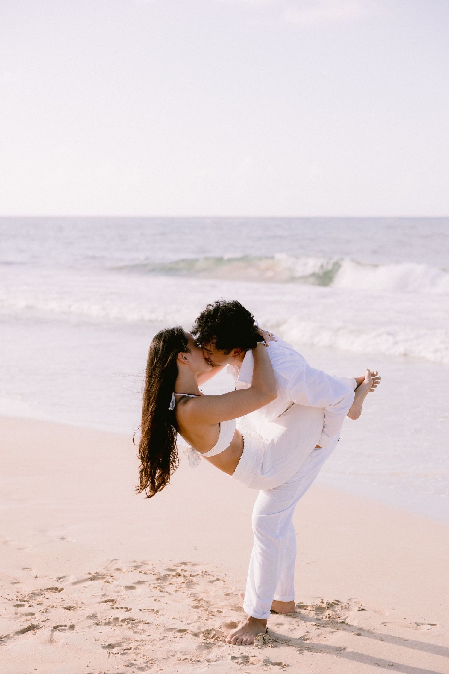 A couple in white outfits sharing romantic moments on a sandy beach.