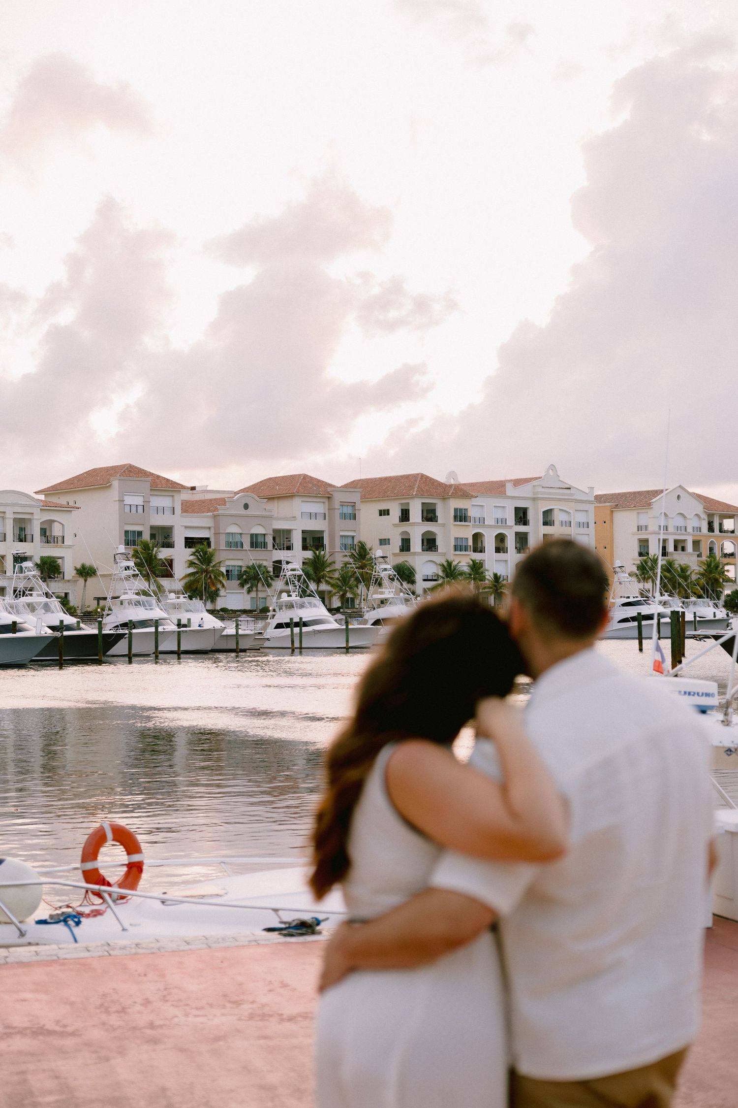Couple embracing at a marina waterfront during sunset with luxury resort buildings and yachts in the background.