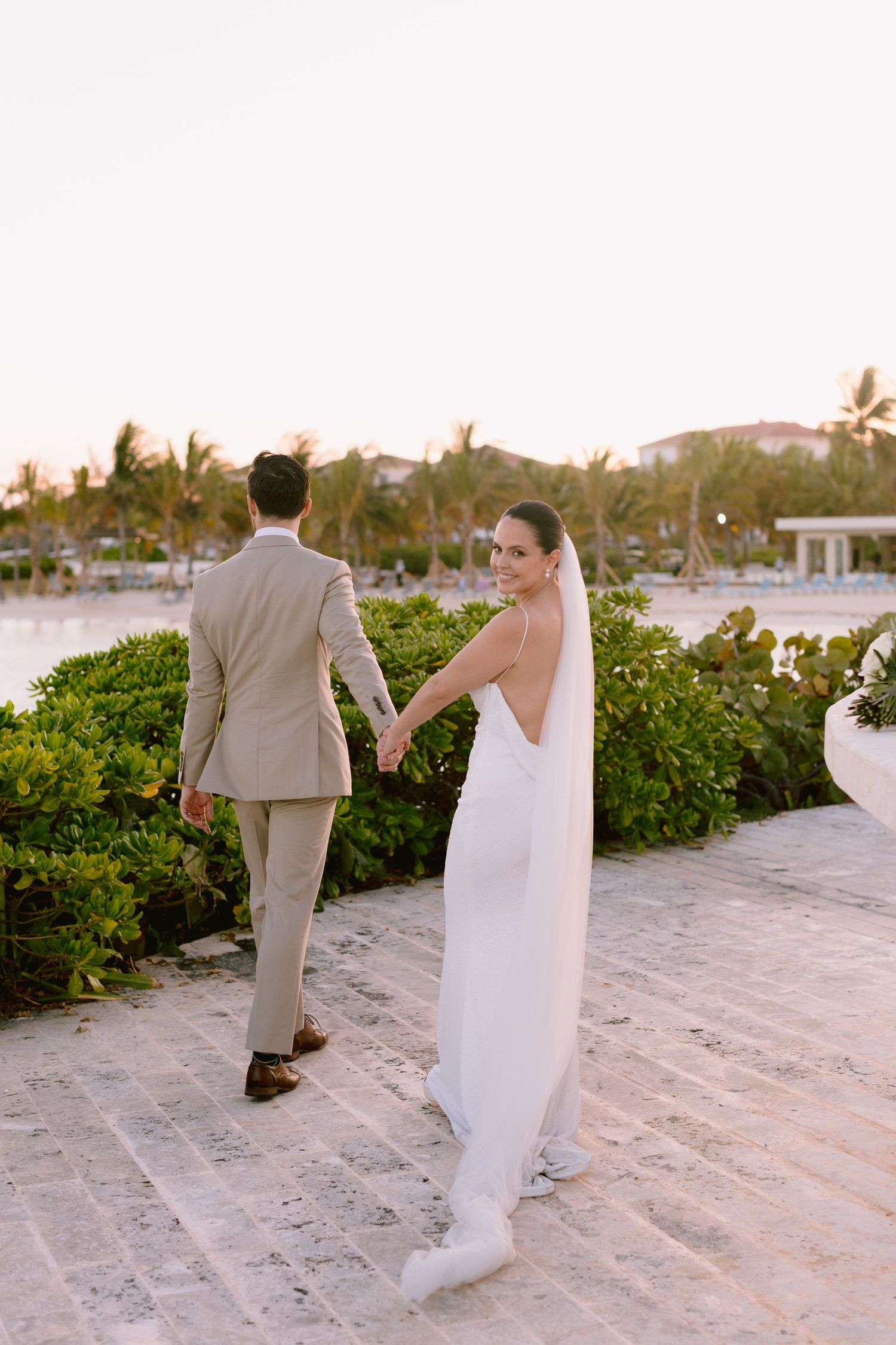 A couple in wedding attire walk together on a sandy beach path lined with tropical greenery at sunset.