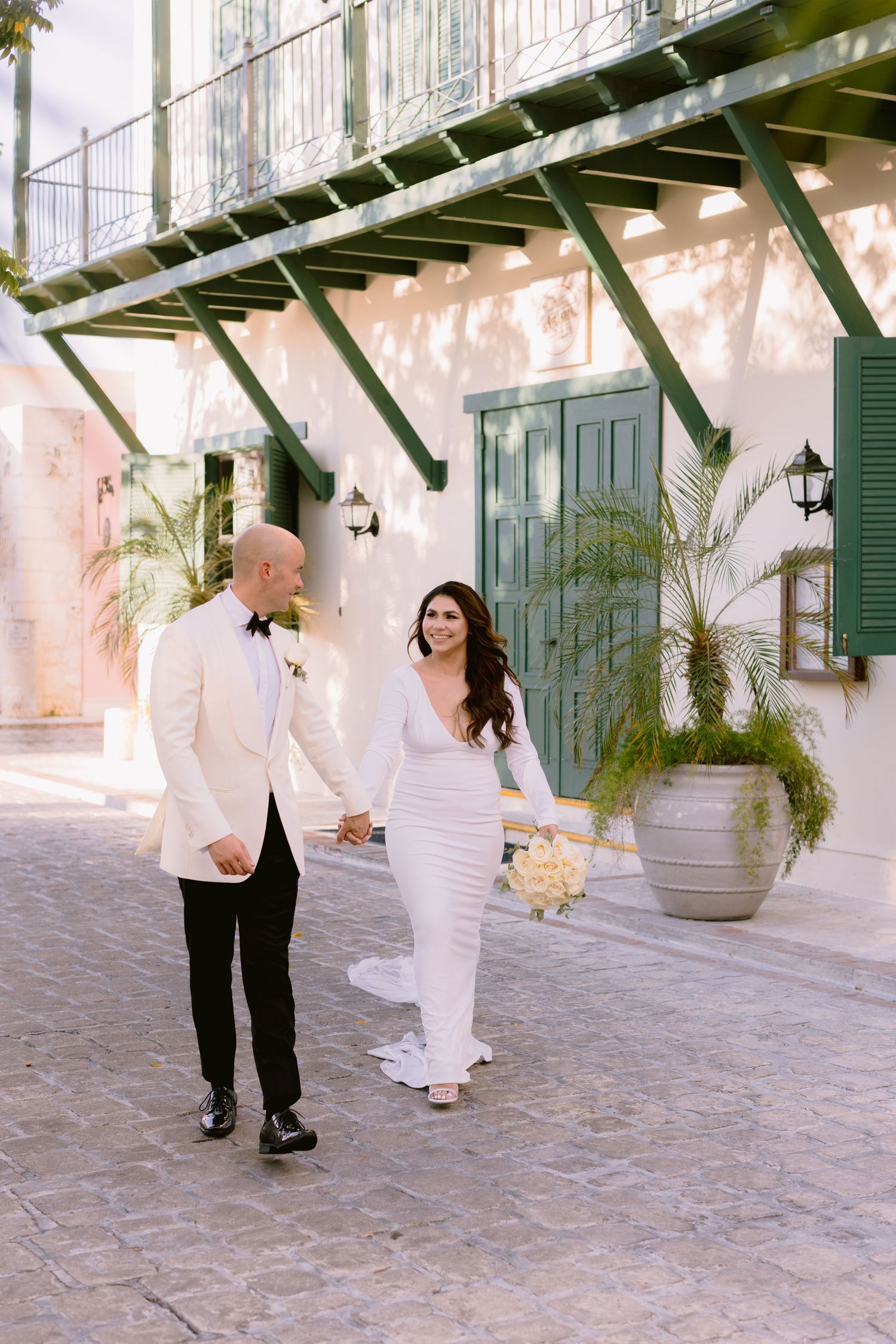 Wedding couple walking along historic courtyard with white architecture and greenery.