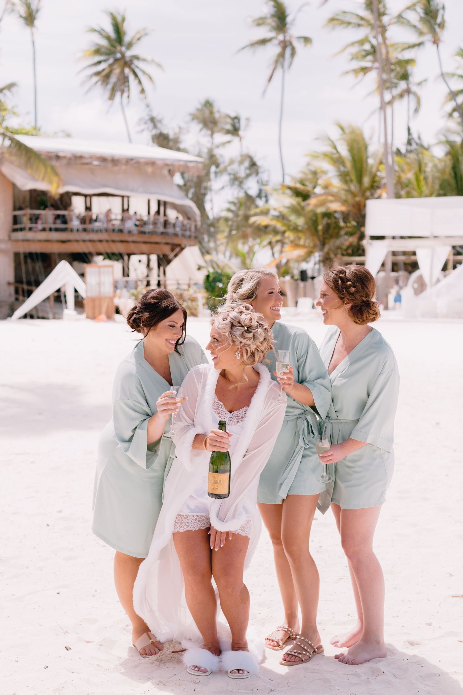 Bridesmaids in matching mint green robes playfully pose on a tropical beach.