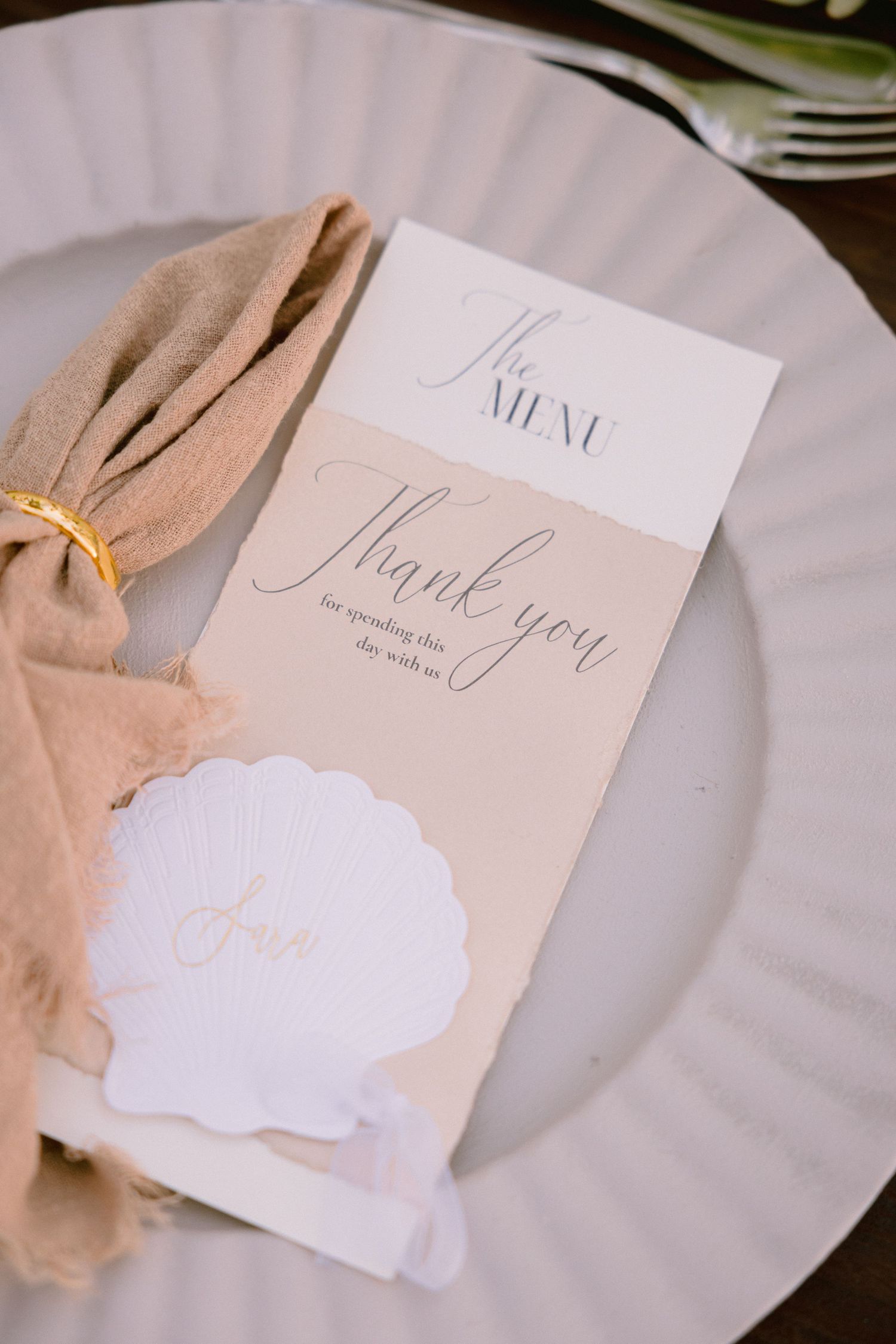 Elegant wedding place setting with menu card, thank you note, seashell name card, and tan linen napkin on white plate.