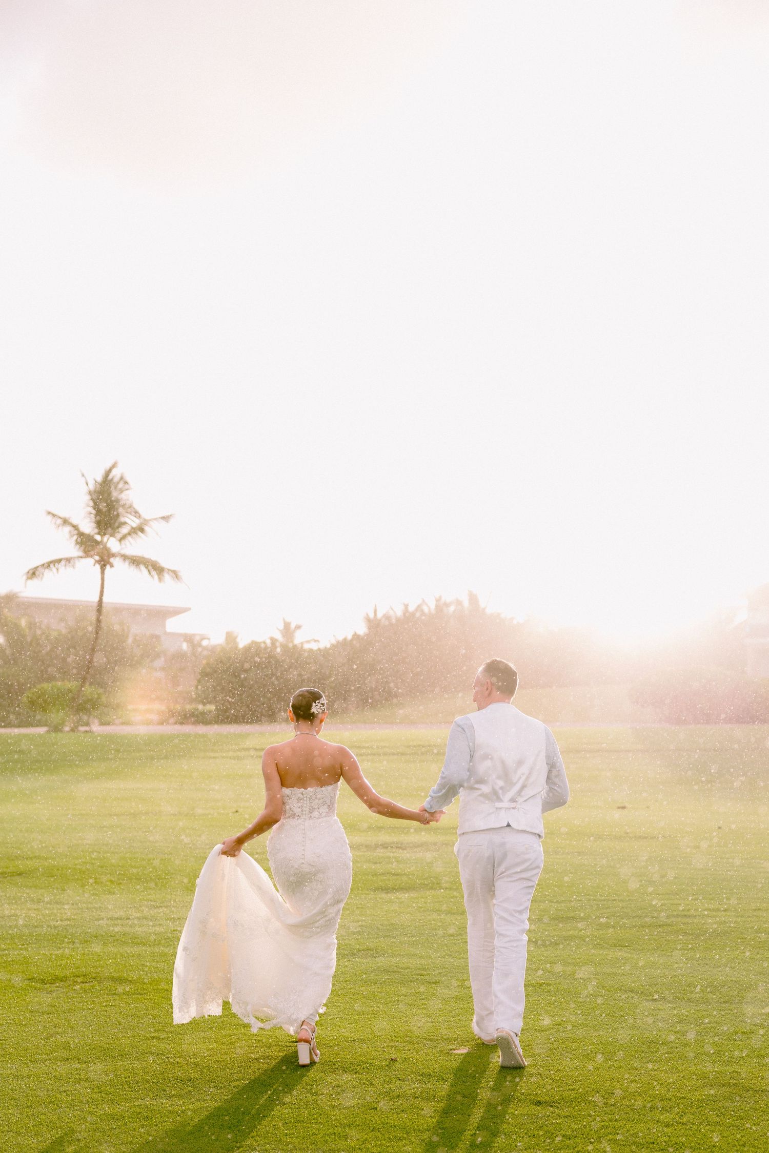 A couple in wedding attire walks hand in hand across a sunlit green lawn with palm trees in the background.