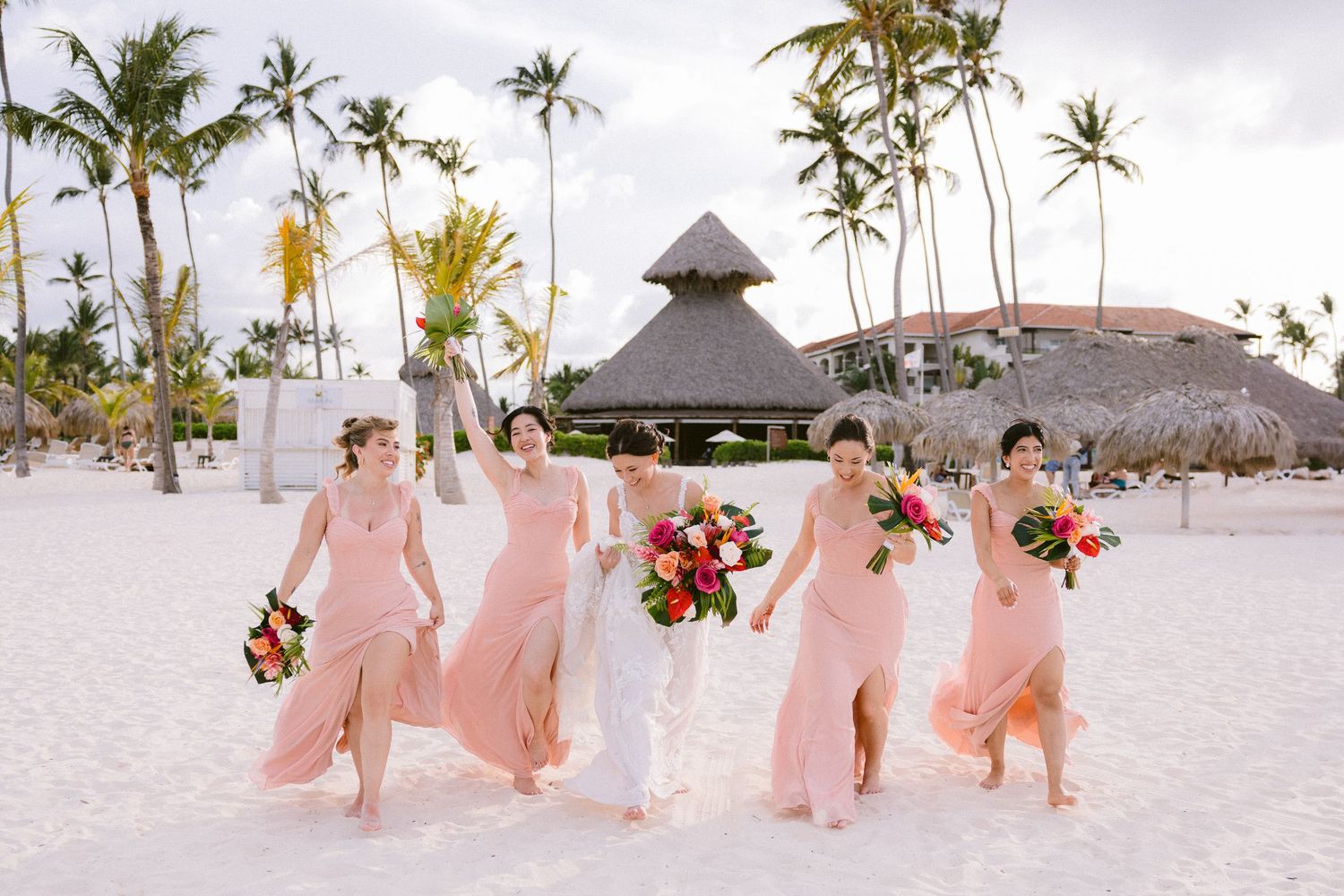 Bridesmaids in blush pink dresses walk along a pristine white sandy beach with palm trees in the background.