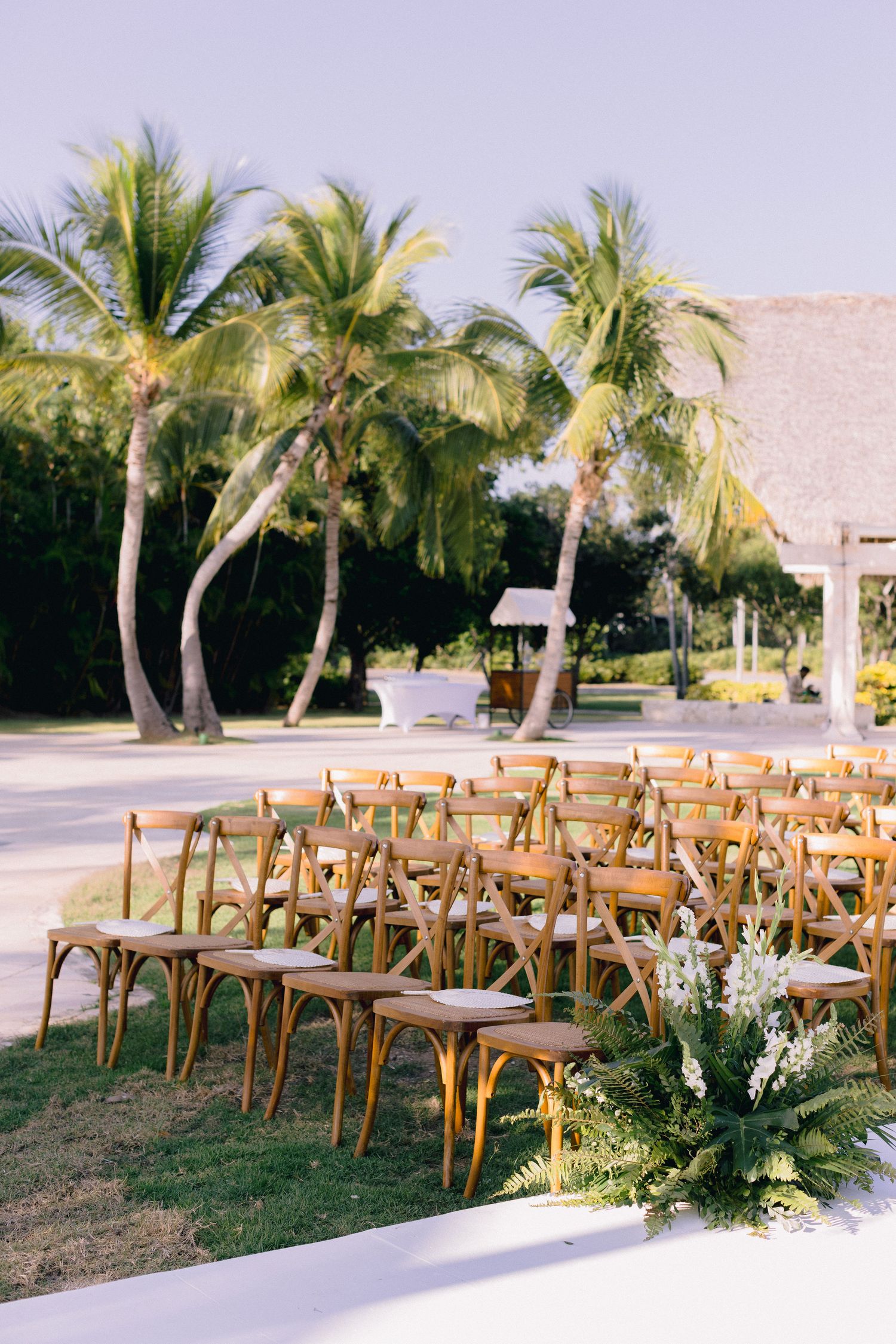 Rustic wooden cross-back chairs arranged in rows for an outdoor beach wedding ceremony with palm trees in the background.