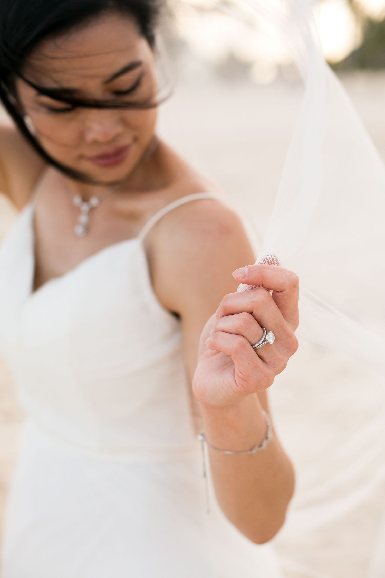 Close-up shot of a bride wearing a delicate bracelet and wedding ring with a soft-focus veil in the background.