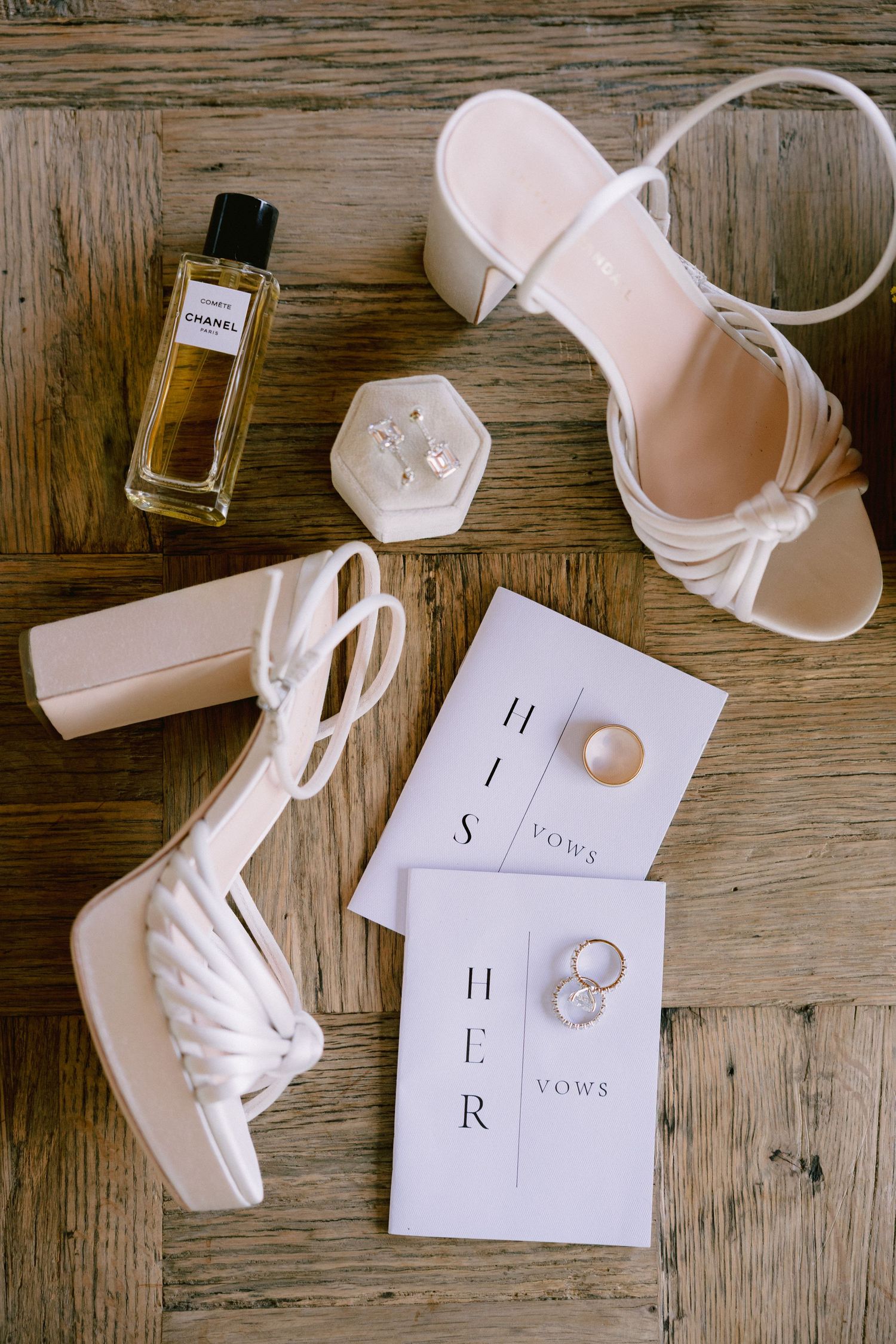 Wedding details flatlay with white bridal shoes, perfume bottle, rings and invitation cards on rustic wooden surface.