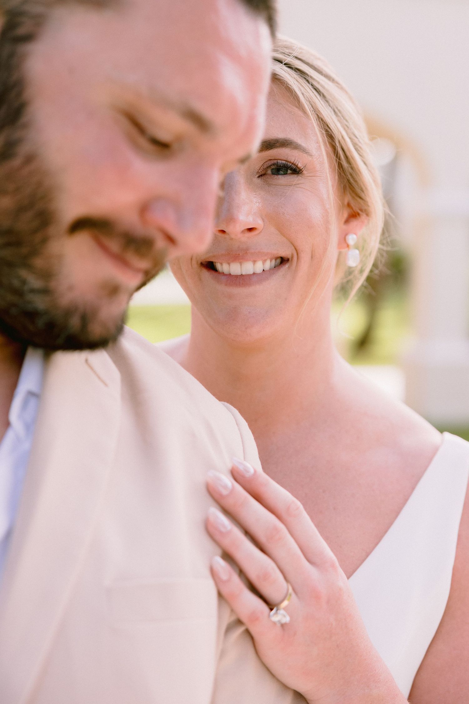 Smiling bride rests hand on groom's shoulder, showcasing diamond engagement ring, in an outdoor wedding photo.
