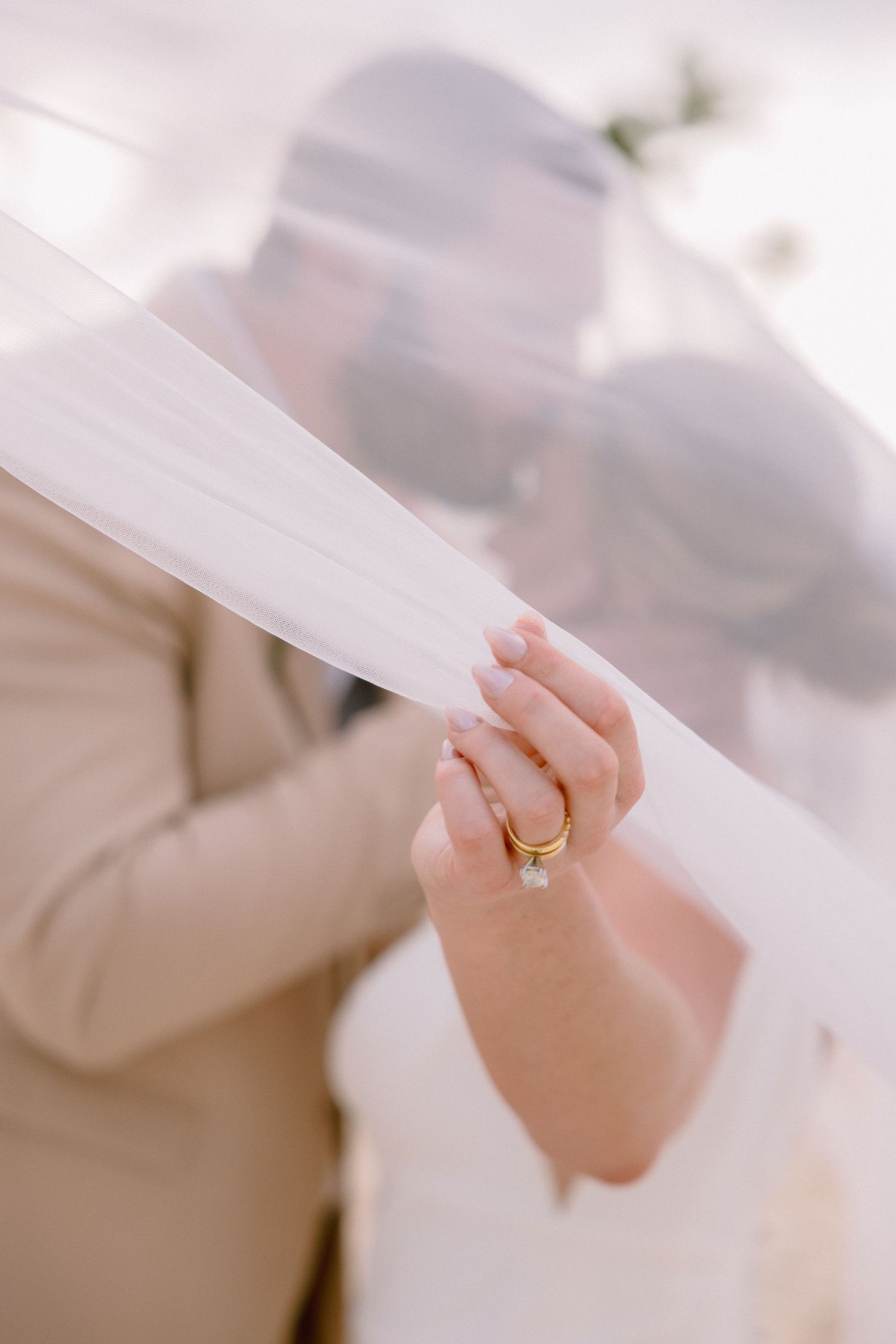 Bride and groom kissing under a flowing white veil, showcasing a gold wedding ring on the bride's hand.