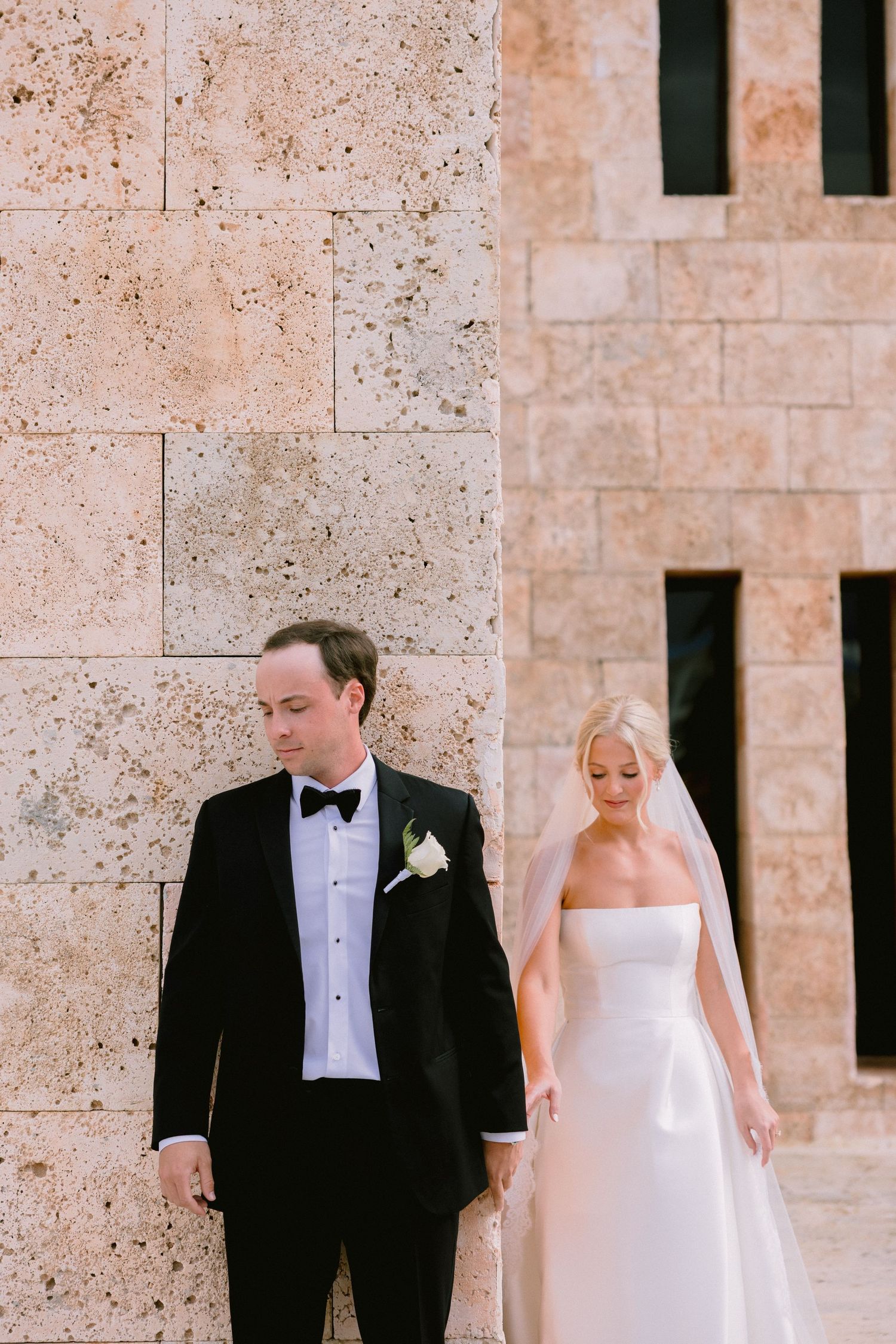 Bride in strapless white gown and veil stands with groom in black tuxedo against a limestone wall.