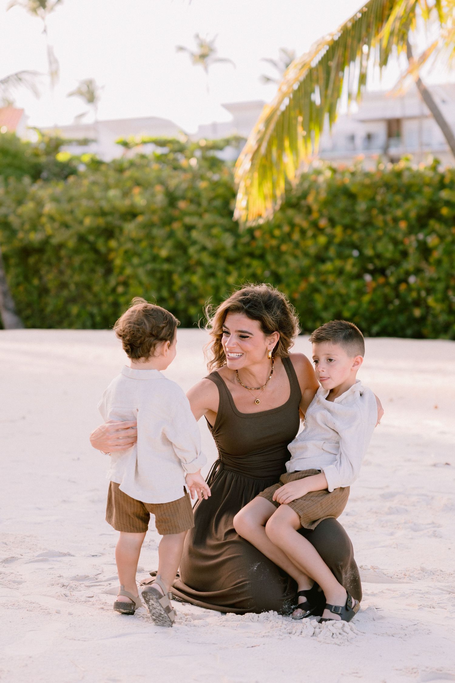 A family relaxes together on a white sandy beach with palm trees and buildings in the background.