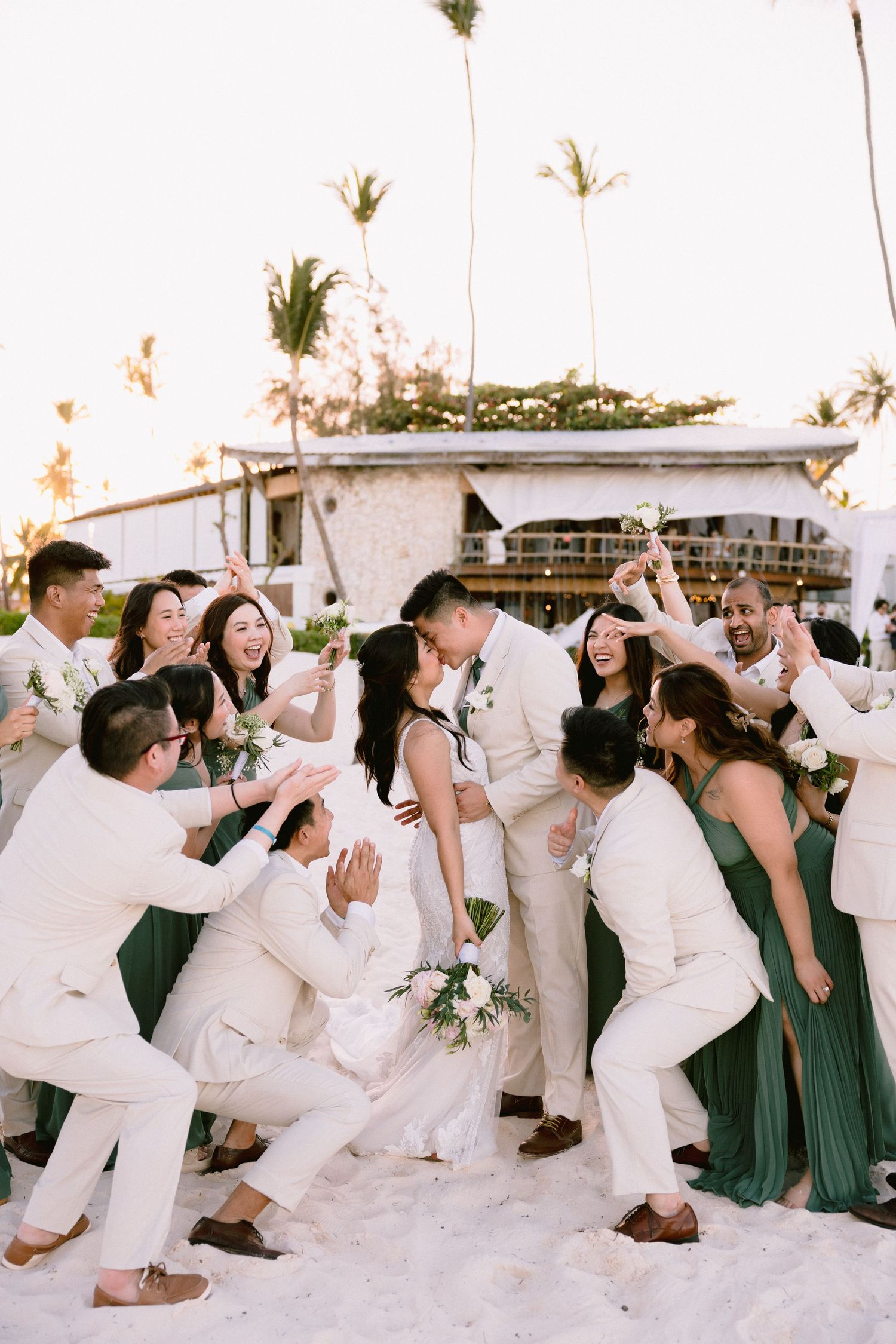 Bride and groom share a kiss surrounded by cheering wedding party in green and white attire on a tropical beach.