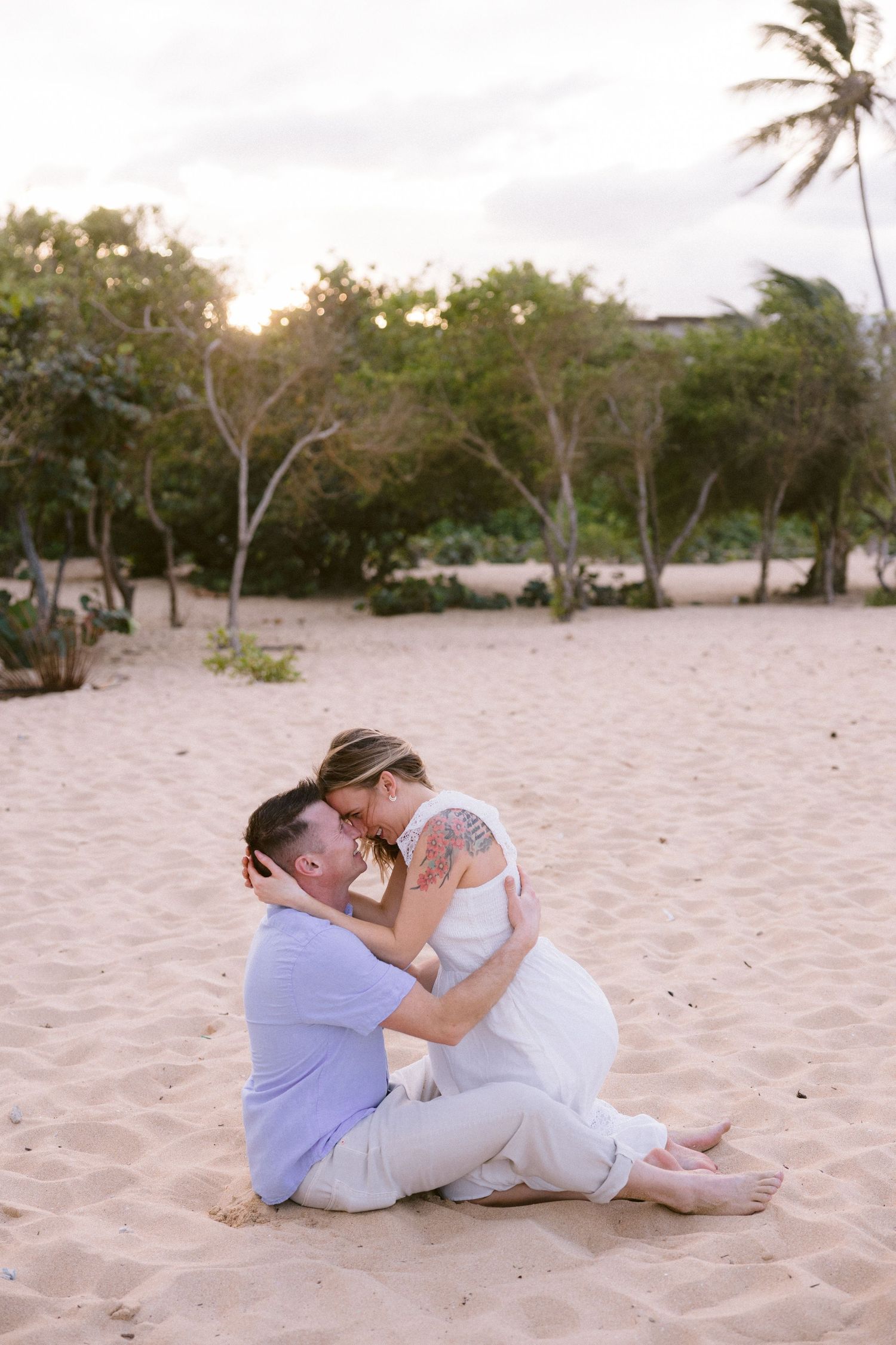 Couple sitting on sandy beach at sunset, sharing a romantic embrace near tropical palm trees and lush greenery.