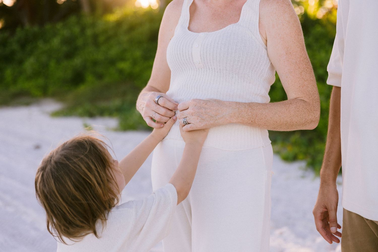 Pregnant woman in white dress with child touching her belly on a sunny beach.