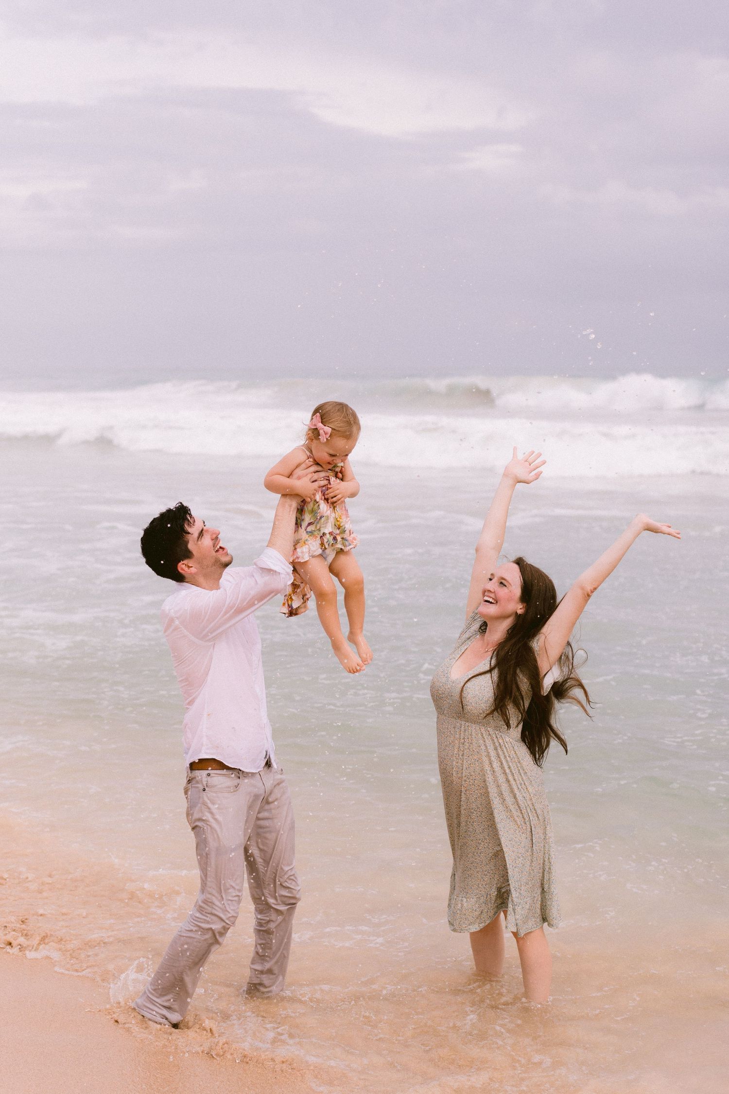 A joyful family plays on a sandy beach, with a man lifting a baby while a woman raises her arms in delight.