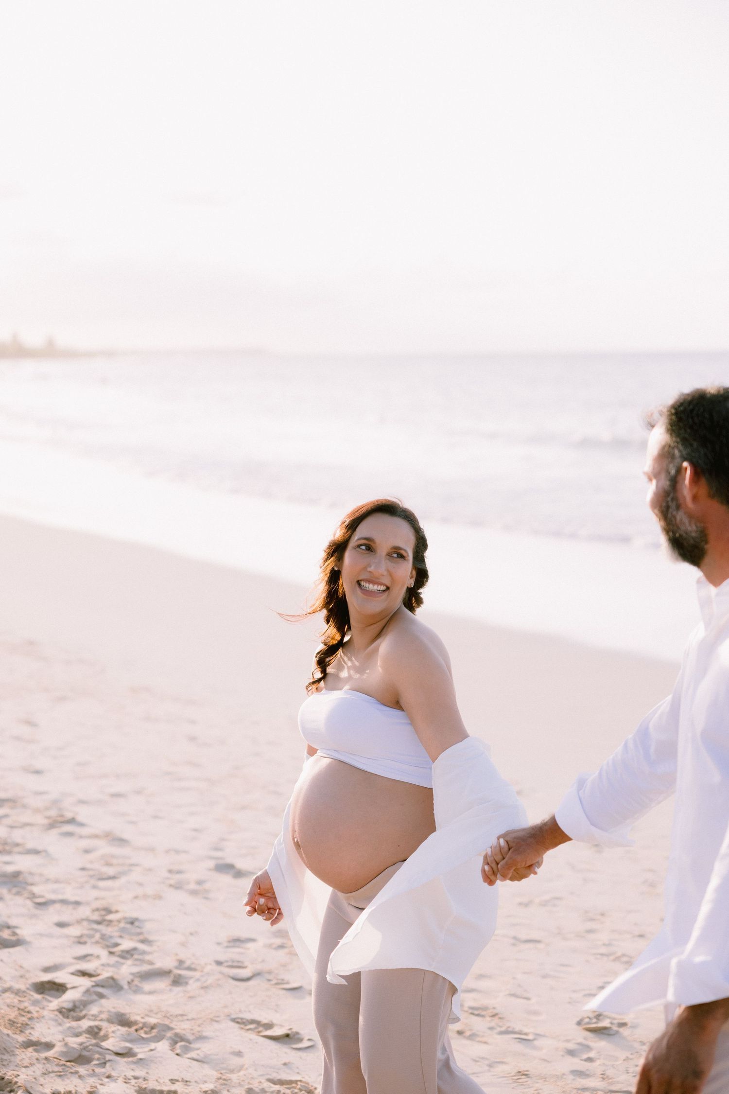 Pregnant woman in white top holds hands with partner on sunny beach, smiling joyfully during maternity photoshoot.