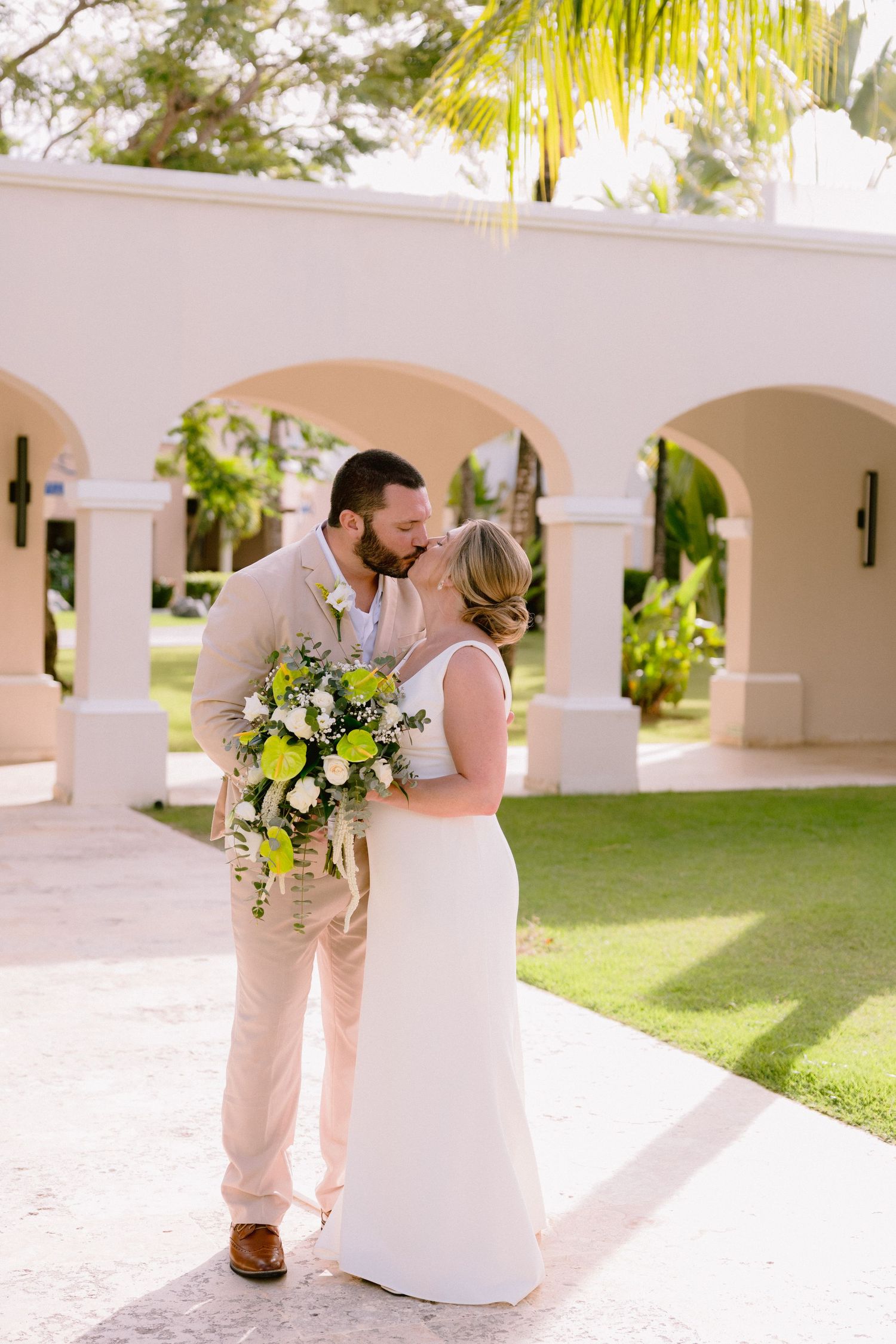 Bride and groom share a kiss outdoors at a tropical wedding venue, bride holding a green and white bouquet.