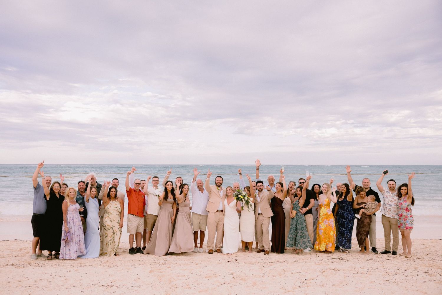 Wedding party celebrating on a beach with hands raised, ocean backdrop, bride in white gown surrounded by guests.