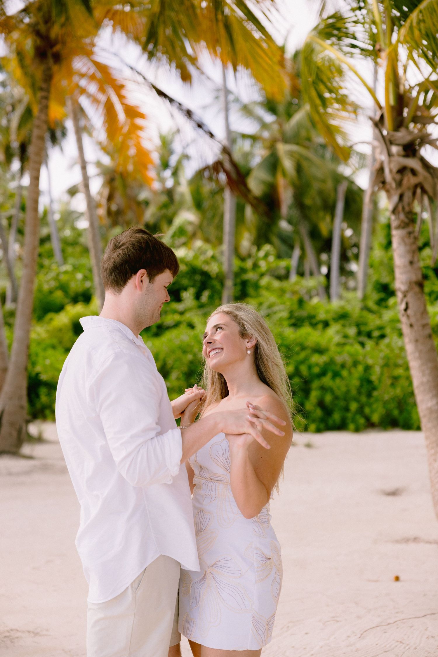 A romantic couple in white clothing dances together on a tropical beach lined with palm trees.