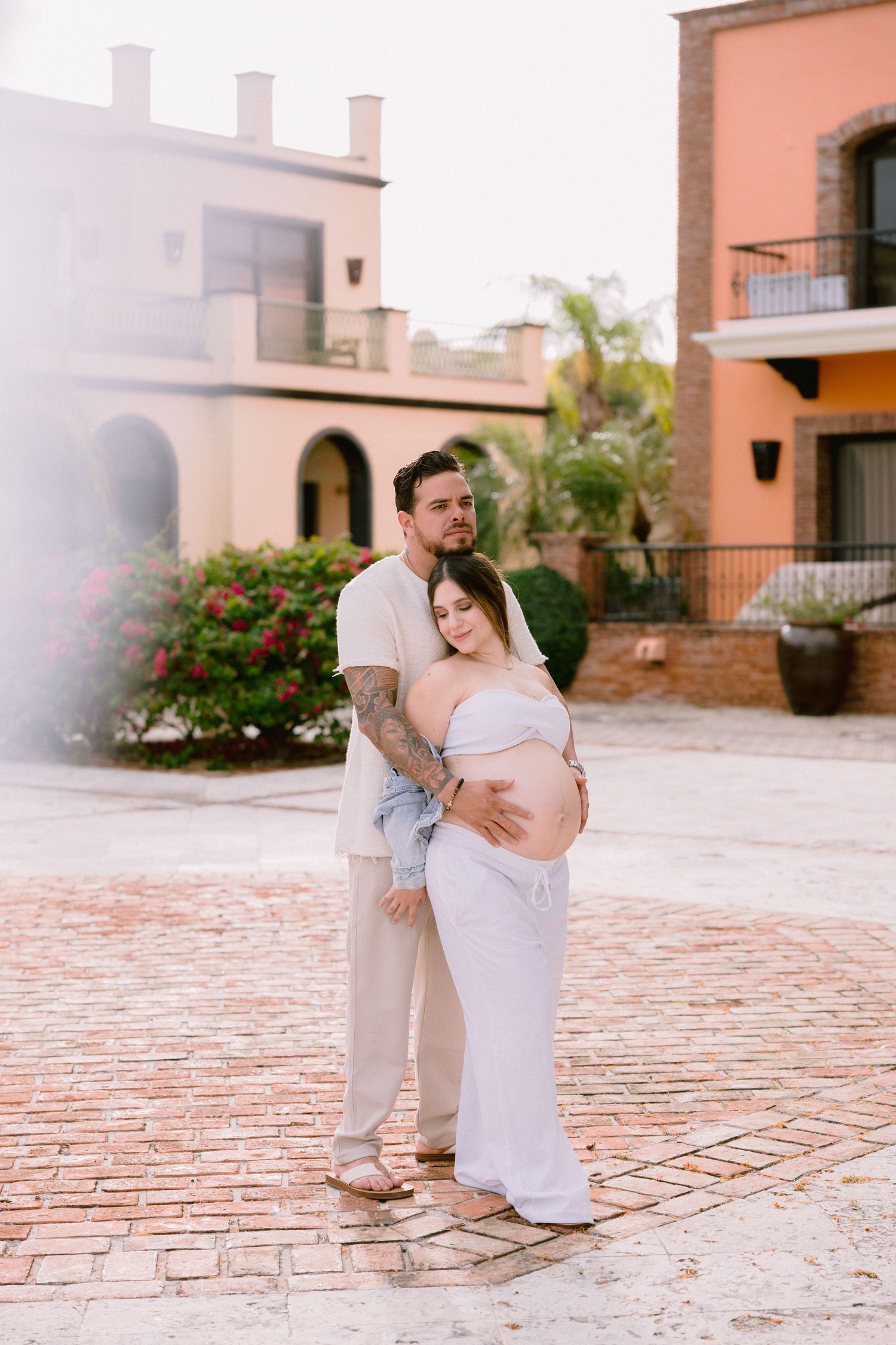 A romantic couple embraces while sitting on a terracotta brick patio near peach-colored Mediterranean style buildings.