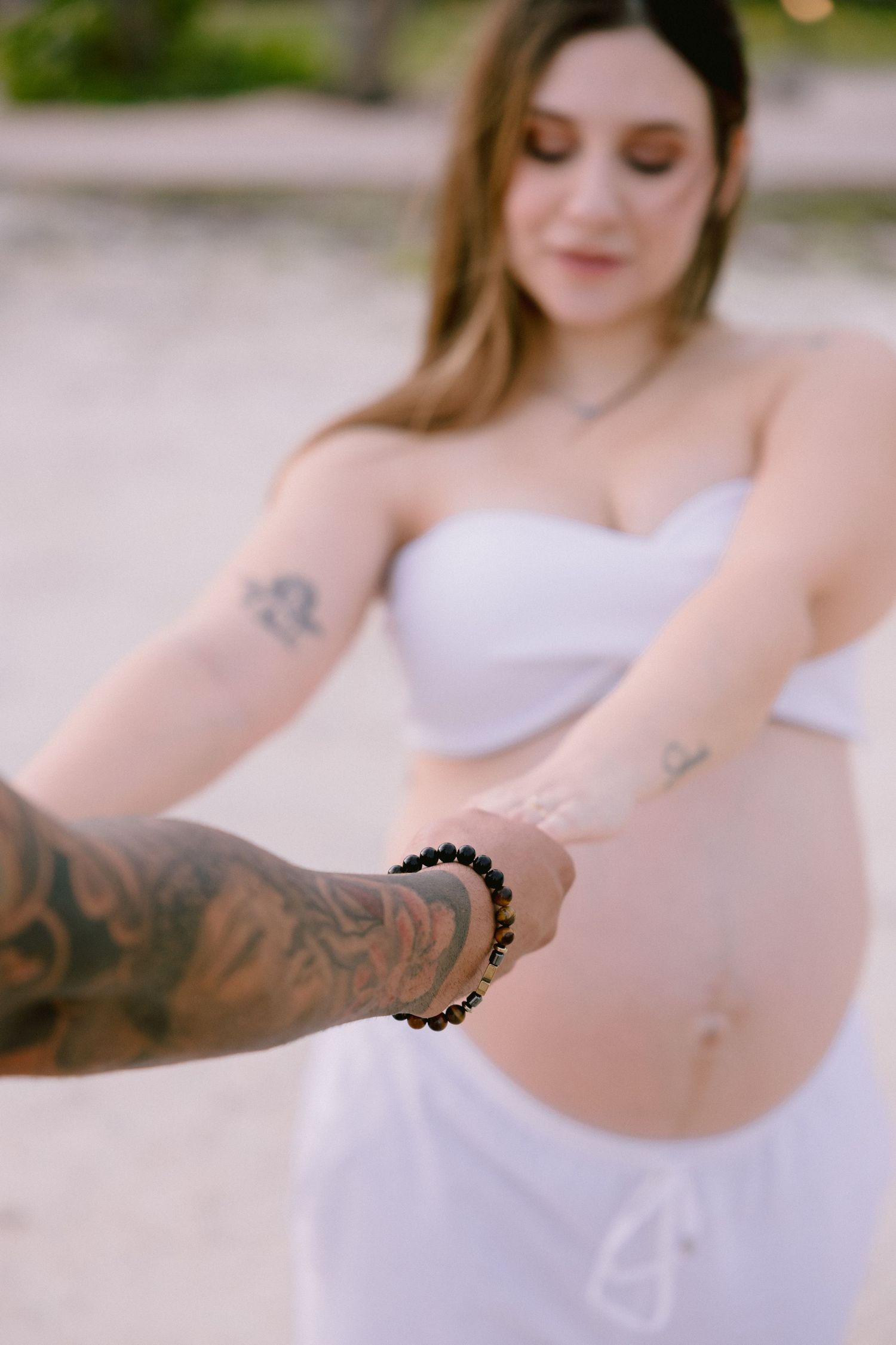 A tattooed hand with beaded bracelet reaches toward figure in white maternity dress against soft blurred background.