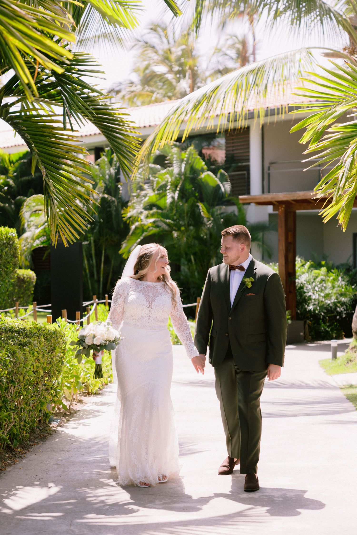 Bride and groom walk hand-in-hand along a tropical resort path surrounded by lush palm trees on their wedding day.