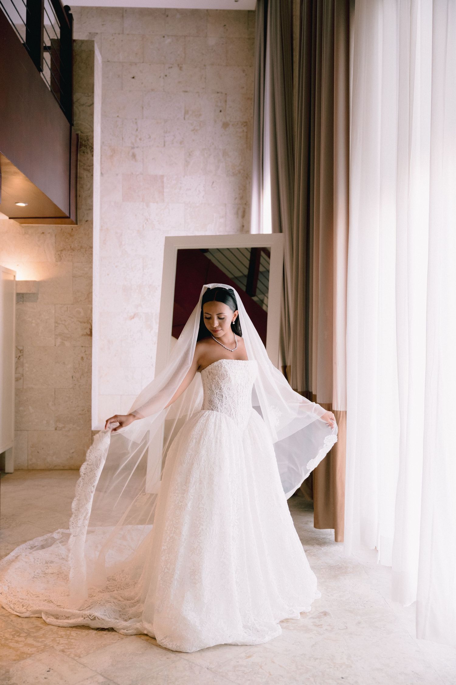 Bride in strapless white ballgown with flowing cathedral veil poses elegantly in a marble-walled venue.