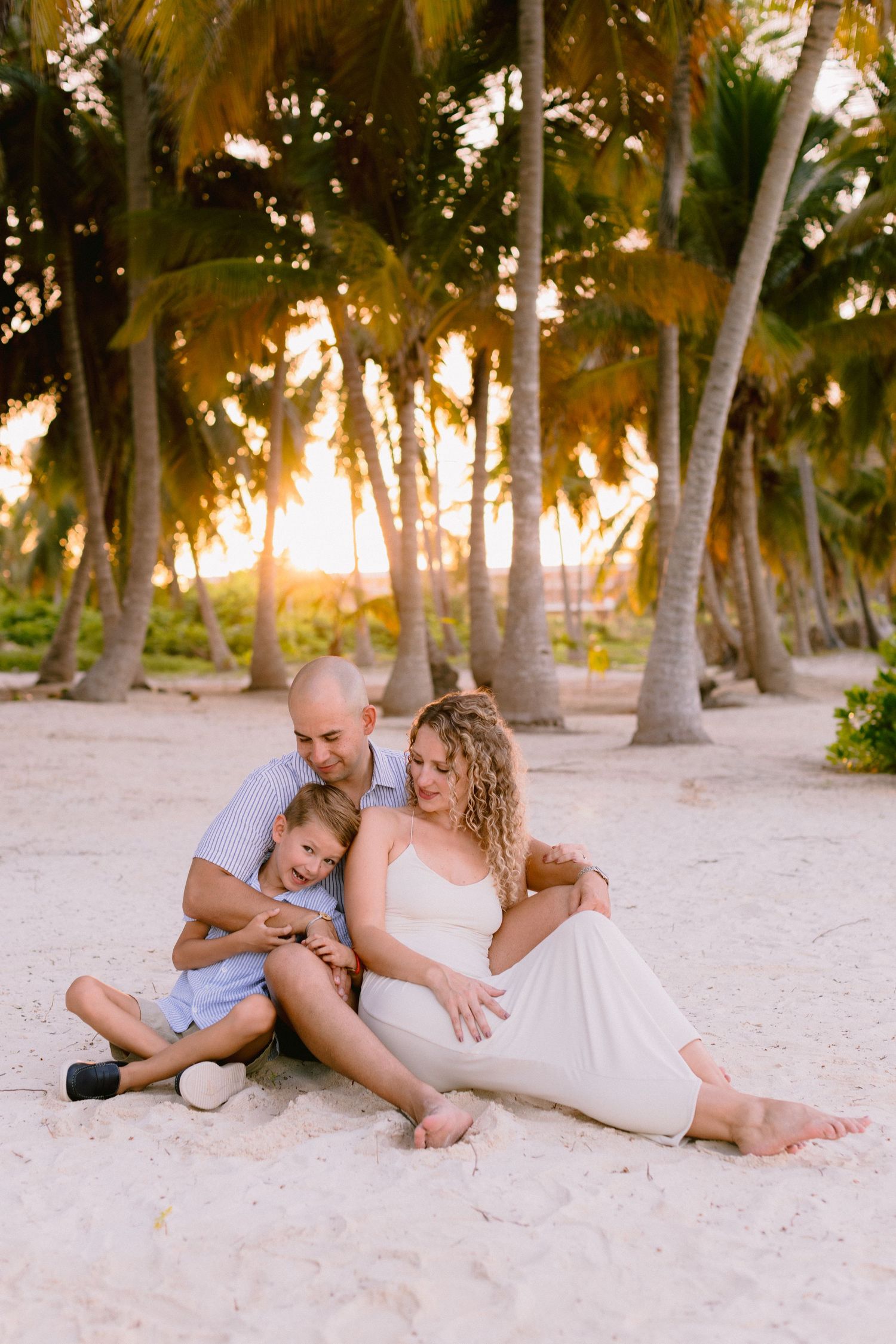 Family of three sitting on sandy beach at golden sunset, surrounded by tropical palm trees, sharing a tender moment.